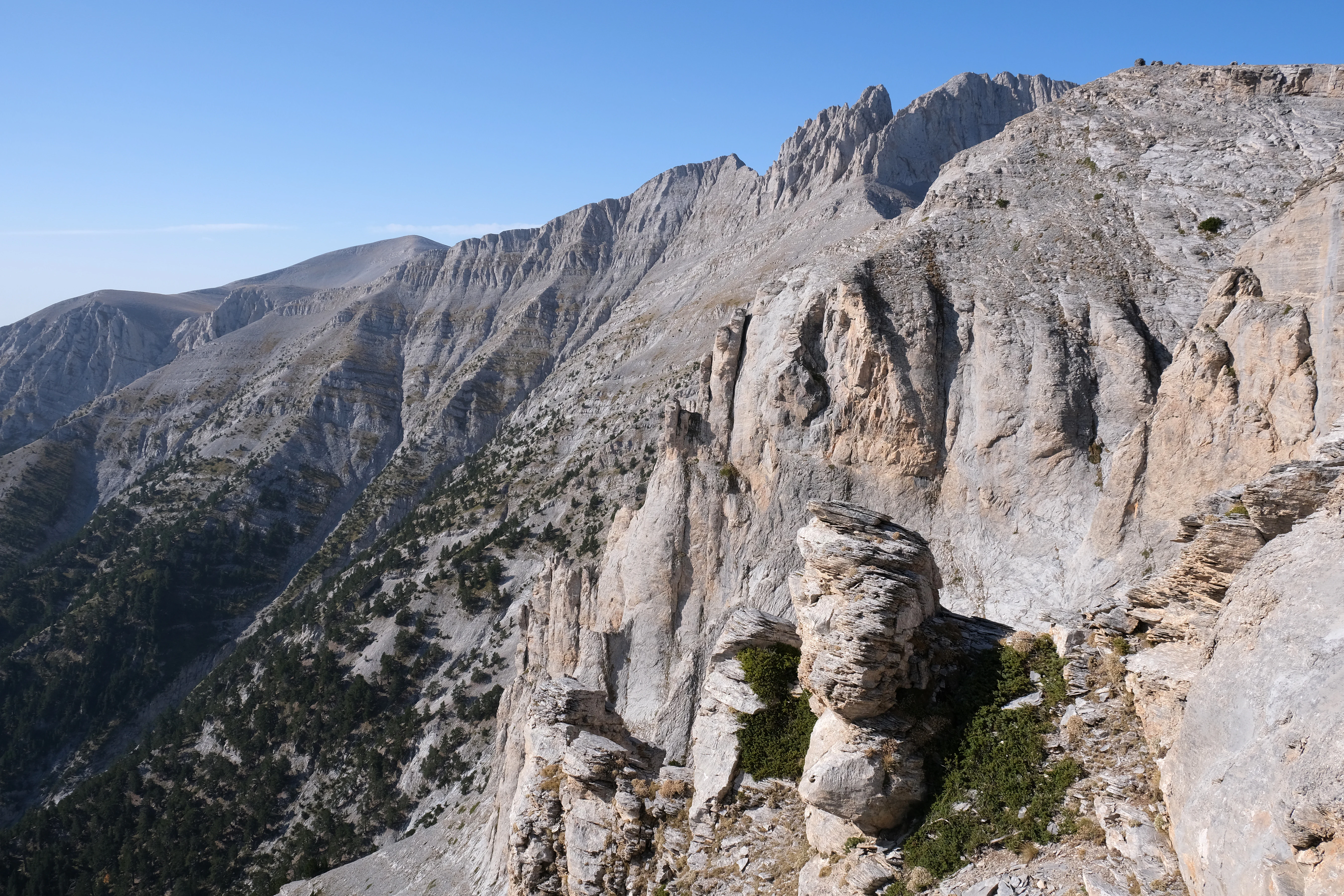 A wide angle view of rocky, light colored mountain peaks and steep cliffs under a clear blue sky in Olympus National Park.