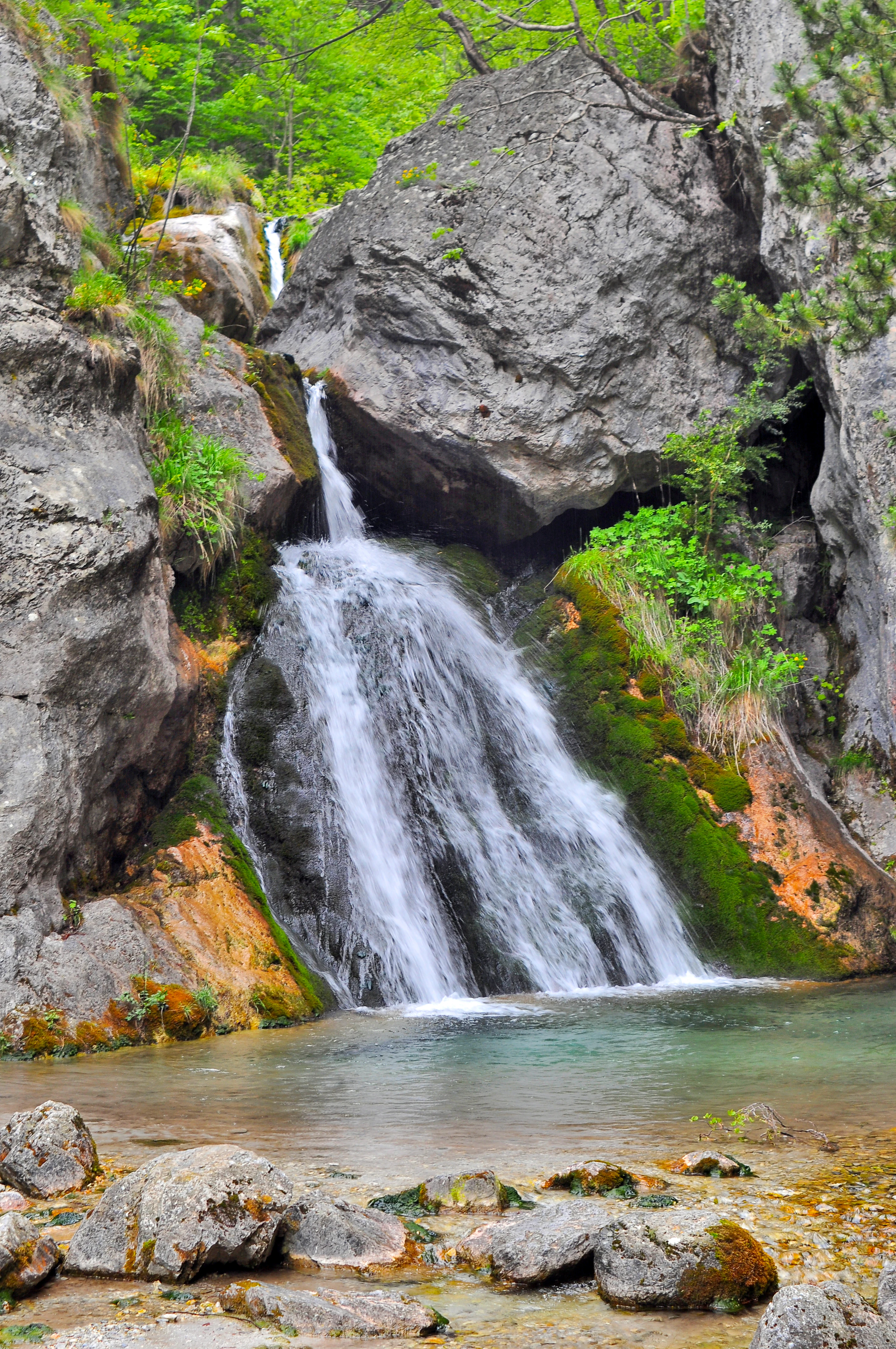 A small waterfall flows down a rocky cliff into a clear mountain pool, with green moss and vegetation covering the surrounding stone.
