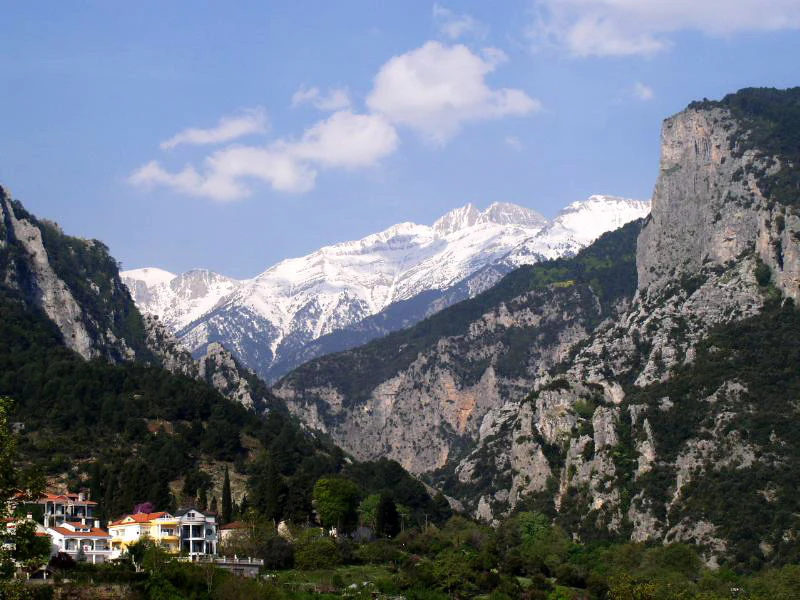A wide scenic view showing the rugged, snow-dusted rocky peaks of Mount Olympus rising behind a Mediterranean town nestled in the foothills.