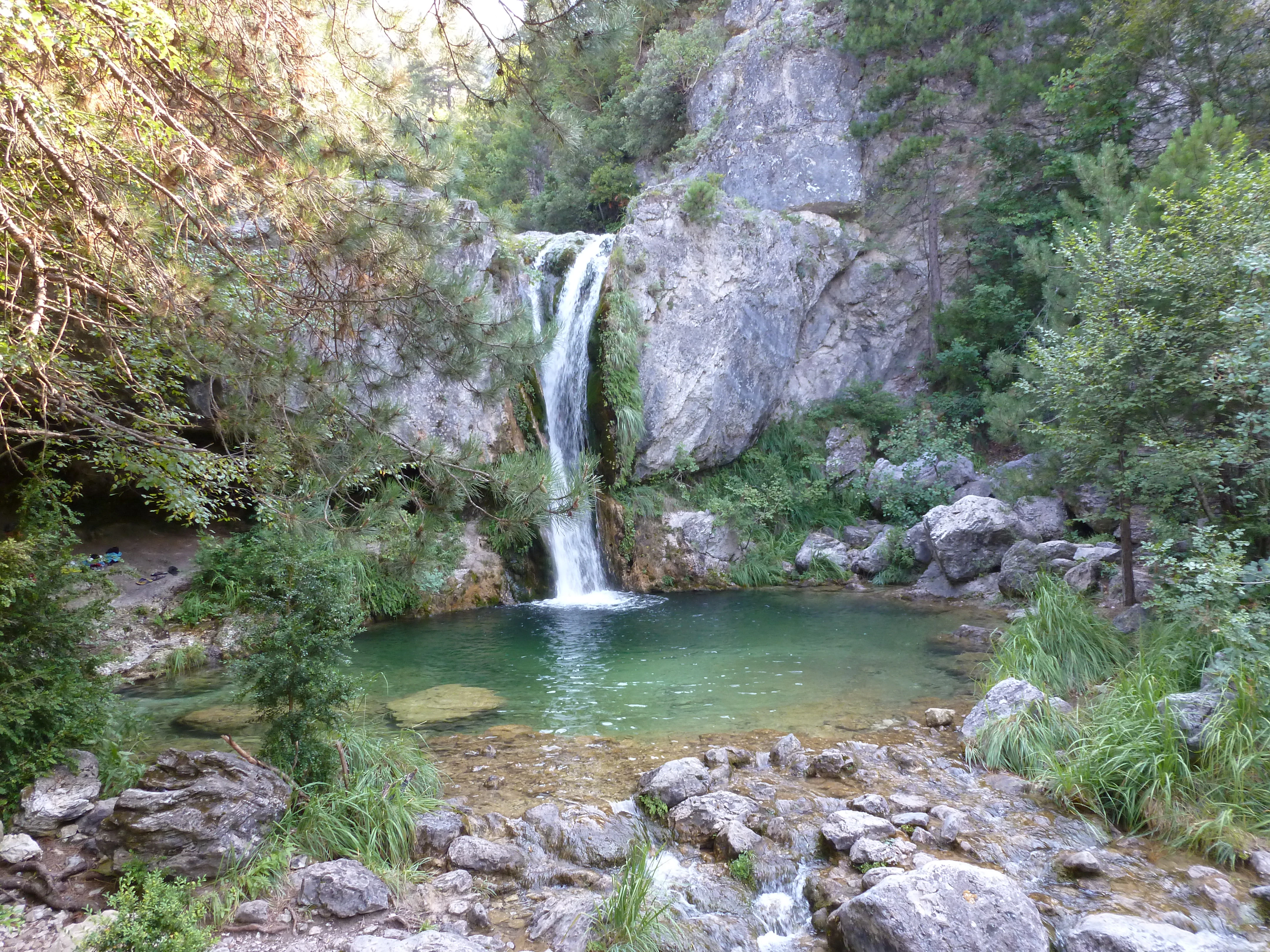 A waterfall cascades down a rocky cliff into a turquoise pool, surrounded by dense forest and stone boulders.