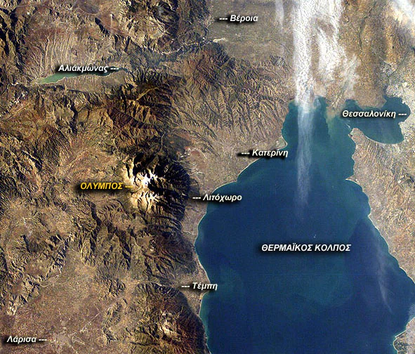An aerial satellite photograph showing the mountainous terrain of Mount Olympus and its proximity to the Thermaic Gulf and surrounding Greek cities, with geographic labels overlaid in Greek.