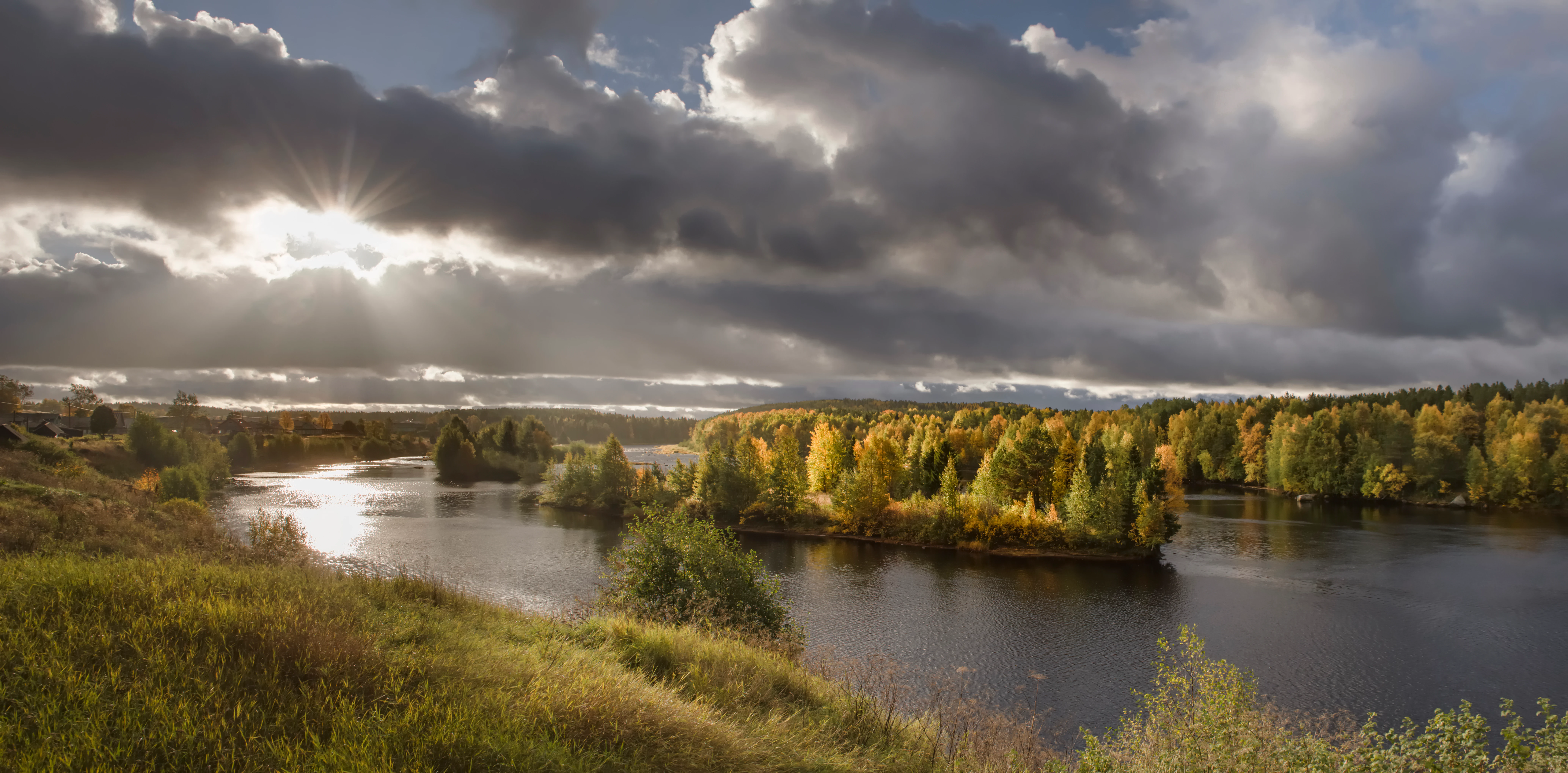 Un río ancho serpentea por un bosque de taiga durante el otoño; los árboles muestran un follaje amarillo y naranja bajo un cielo dramático y nublado con rayos de sol que se abren paso.