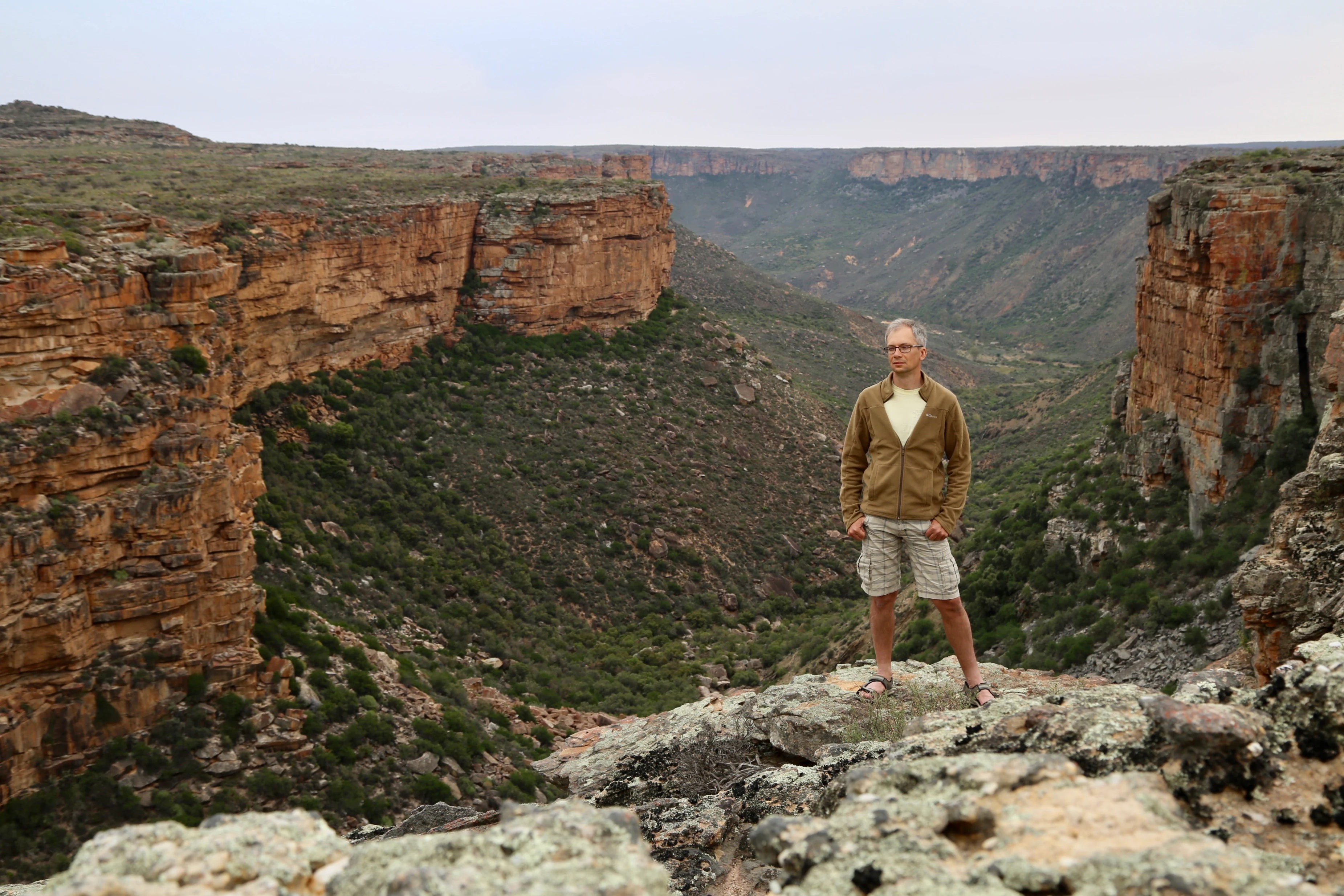 A man standing on a rocky cliff overlooking a deep canyon with rugged walls and sparse vegetation.