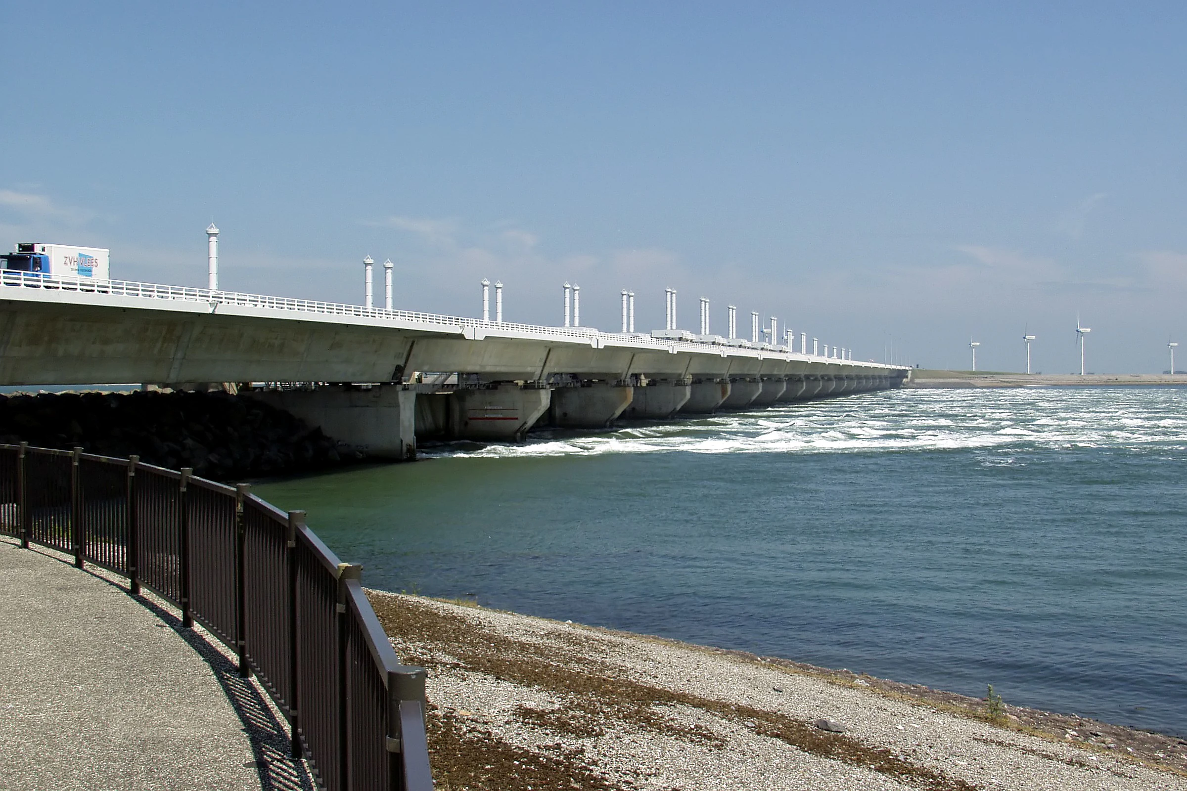 Paved pathway with metal railing leading to a large concrete storm surge barrier spanning a waterway under a clear blue sky