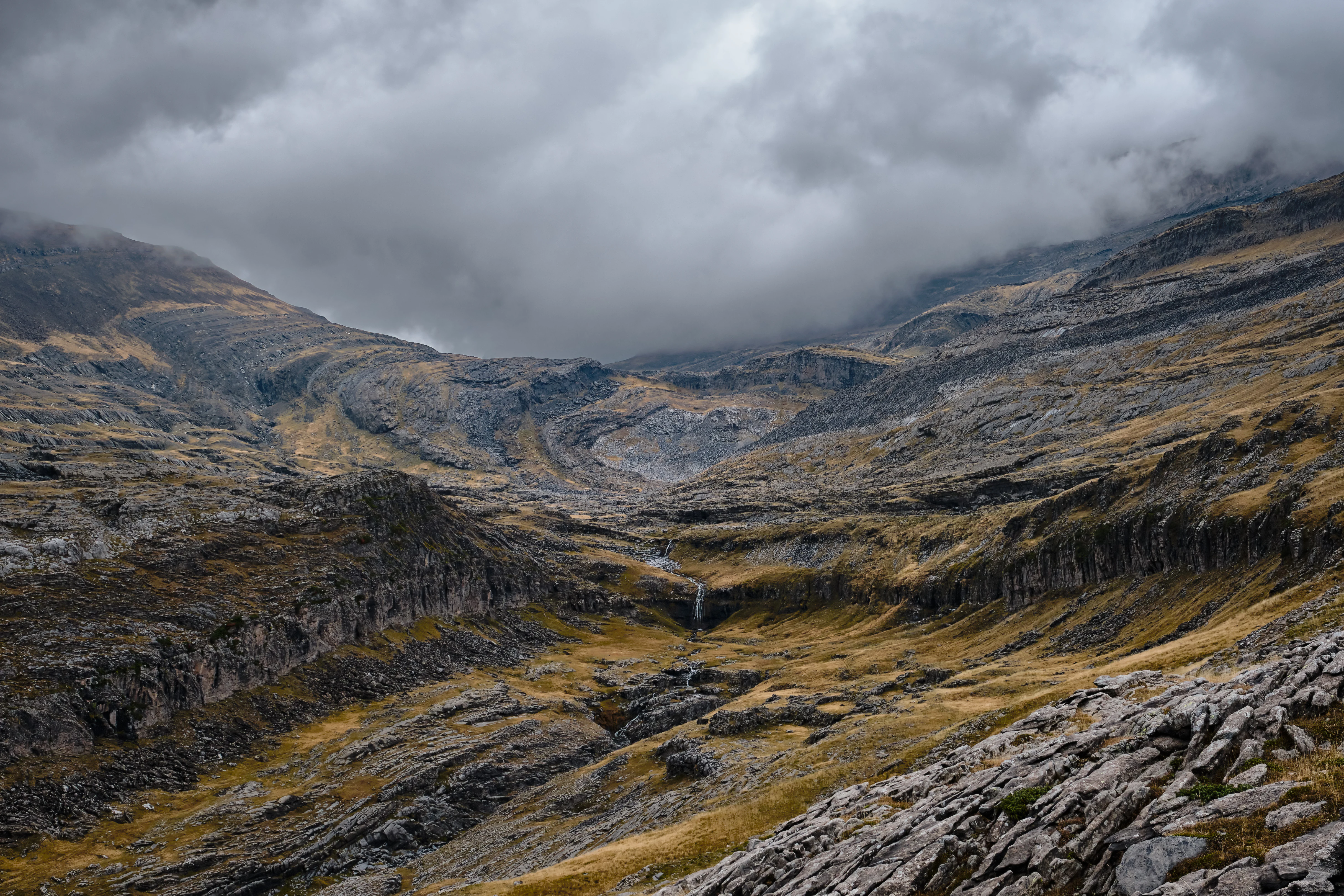 Wide mountain landscape showing a cirque with rocky slopes, patches of grass, and a waterfall under cloudy skies