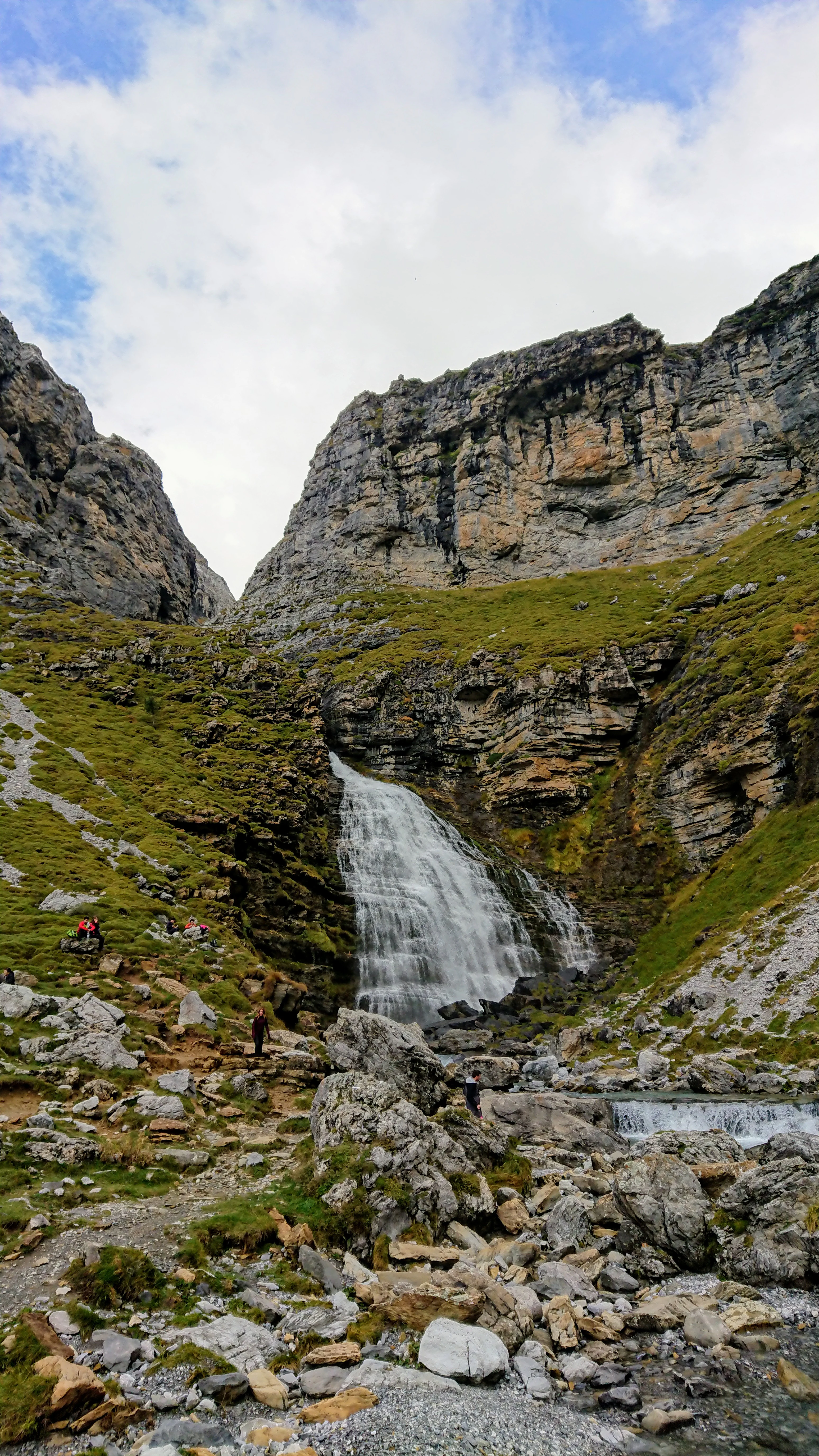 Waterfall cascading down rocky cliffs surrounded by green slopes and boulders with hikers near the base
