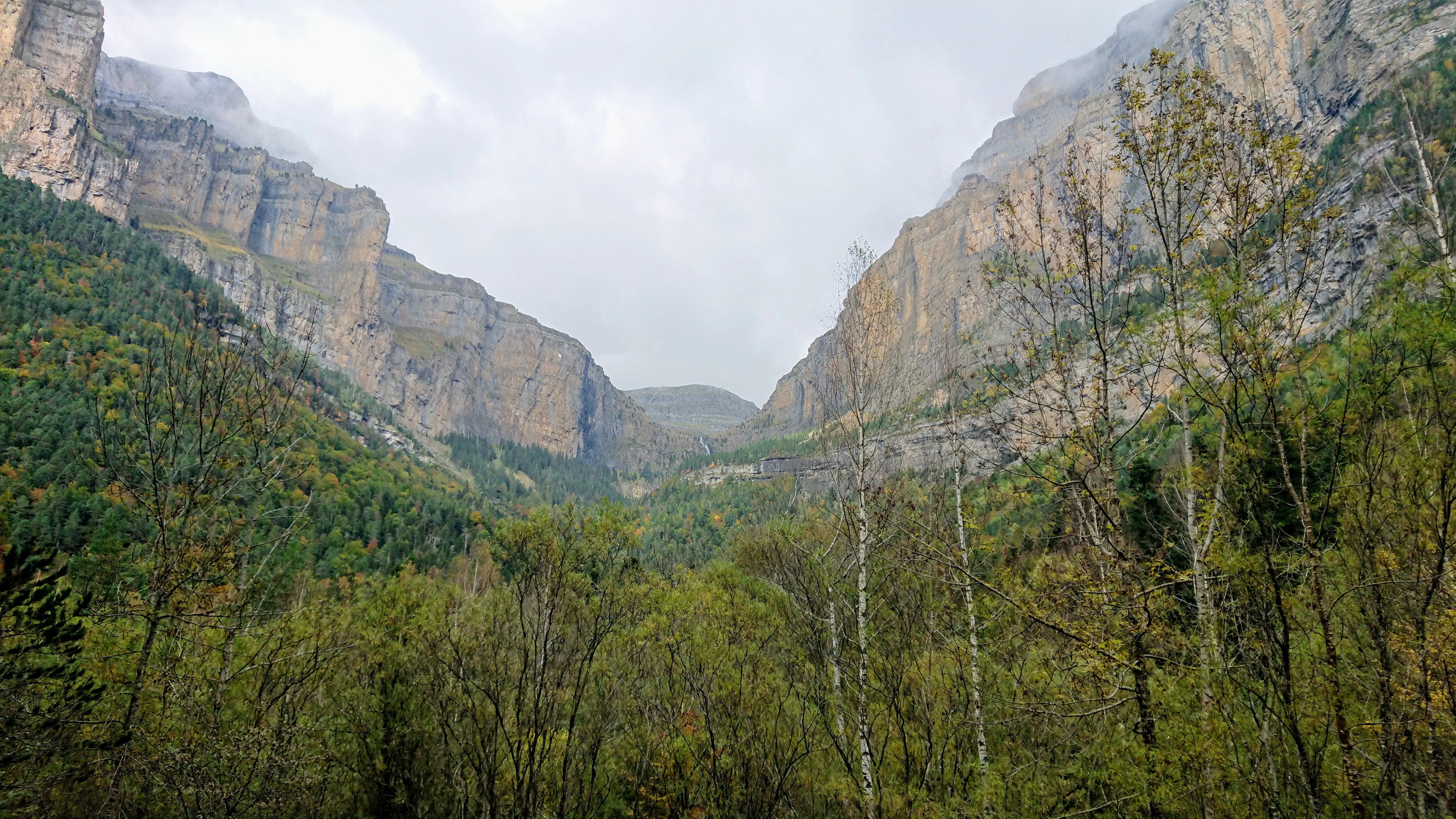 Mountainous landscape featuring a glacial cirque with steep rocky cliffs, forested slopes, and a valley extending into the distance under an overcast sky