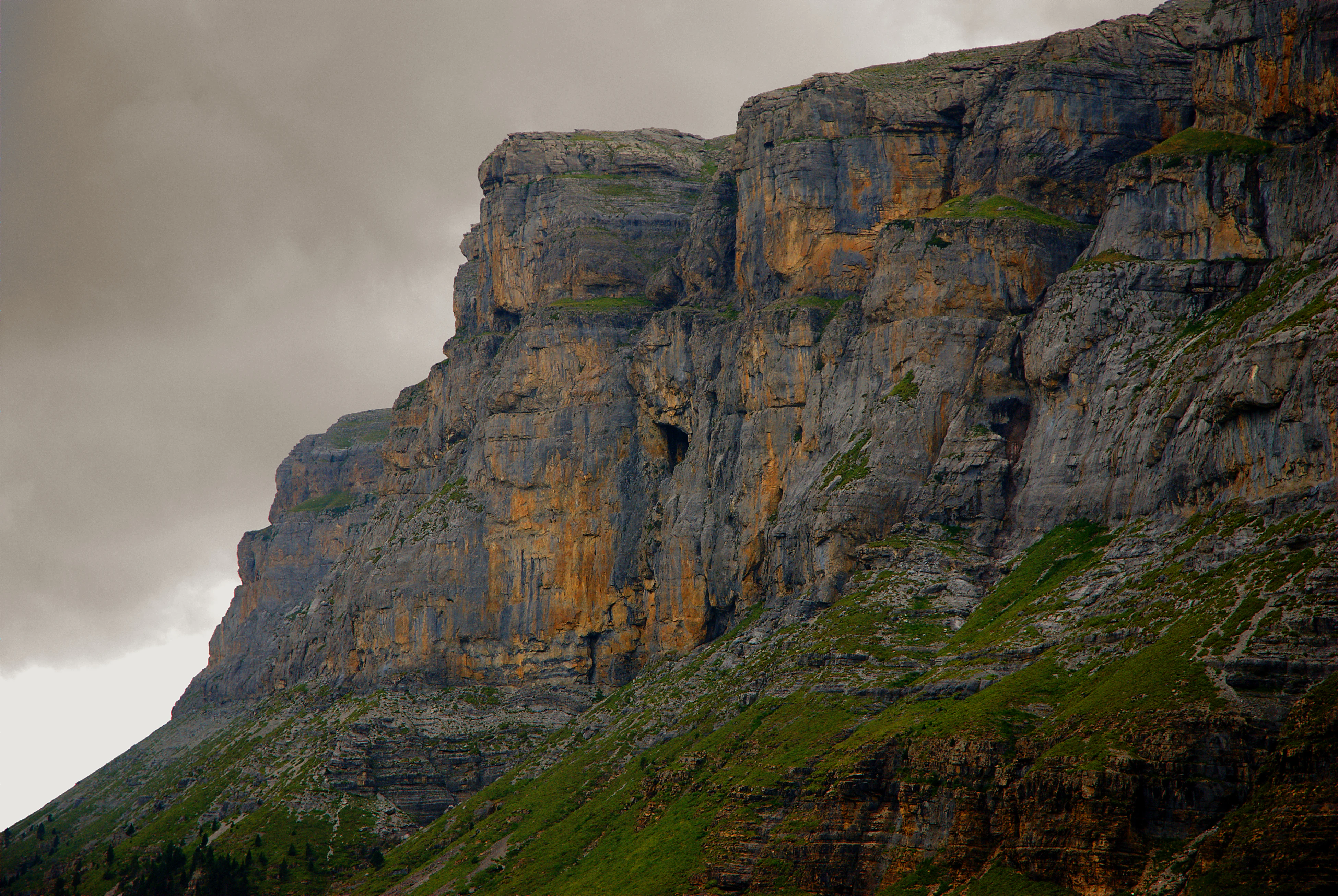 Steep rocky mountain cliffs with layered rock formations and patches of green vegetation under an overcast sky