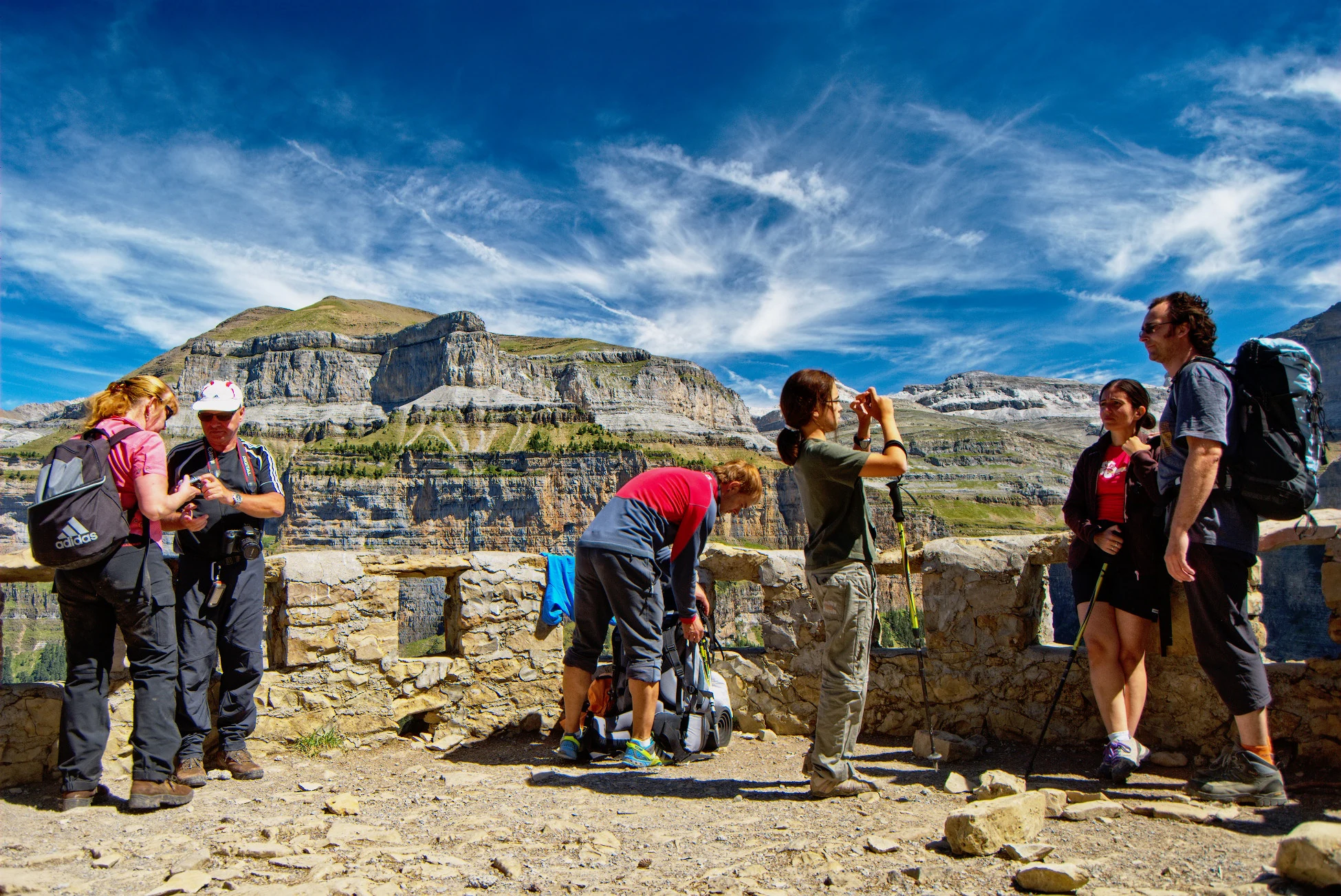 Group of hikers with backpacks and trekking poles at stone viewing platform with mountain range in background