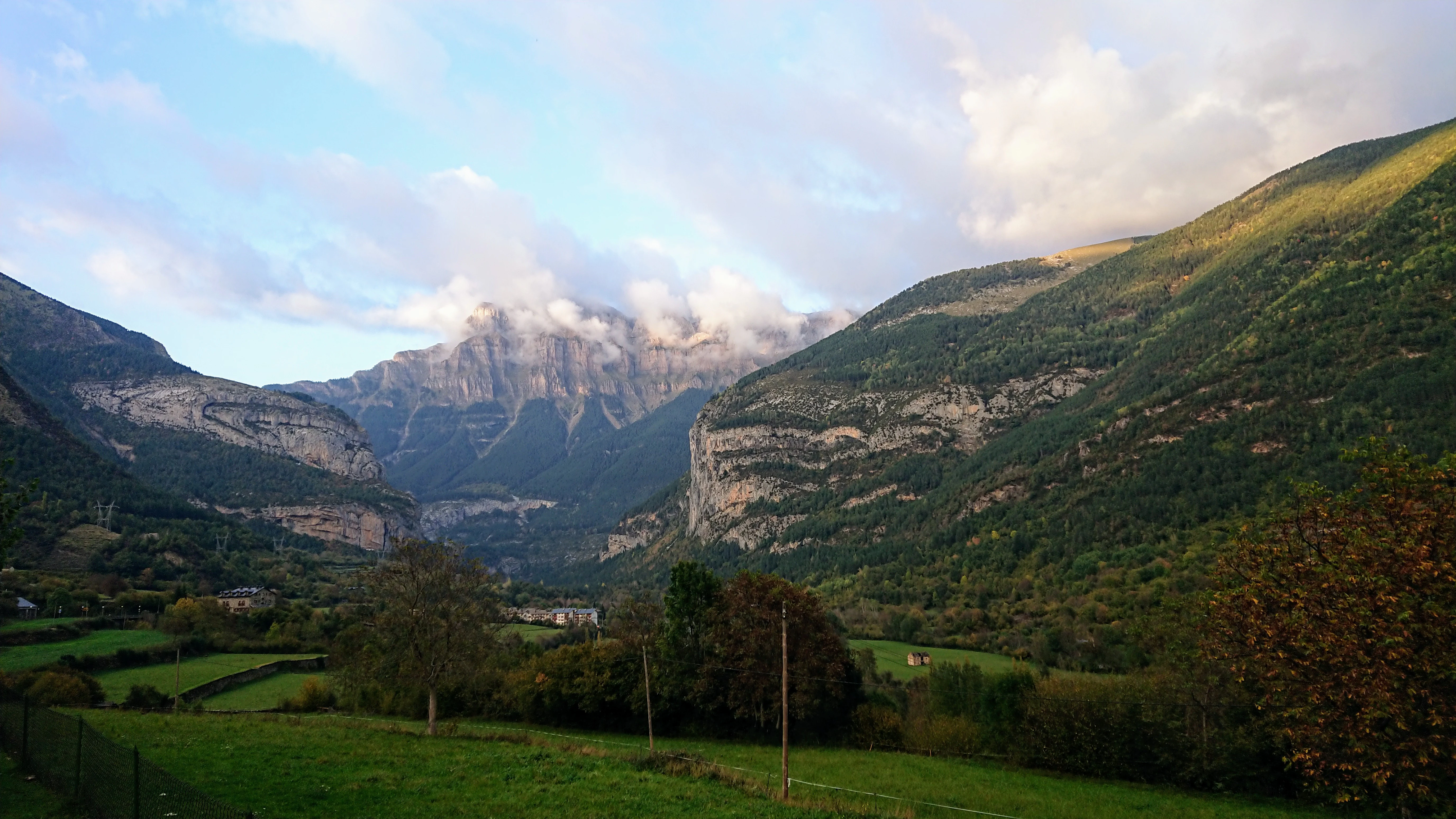 Landscape view of Ordesa Valley entrance with green fields, trees, mountains, and partly cloudy sky
