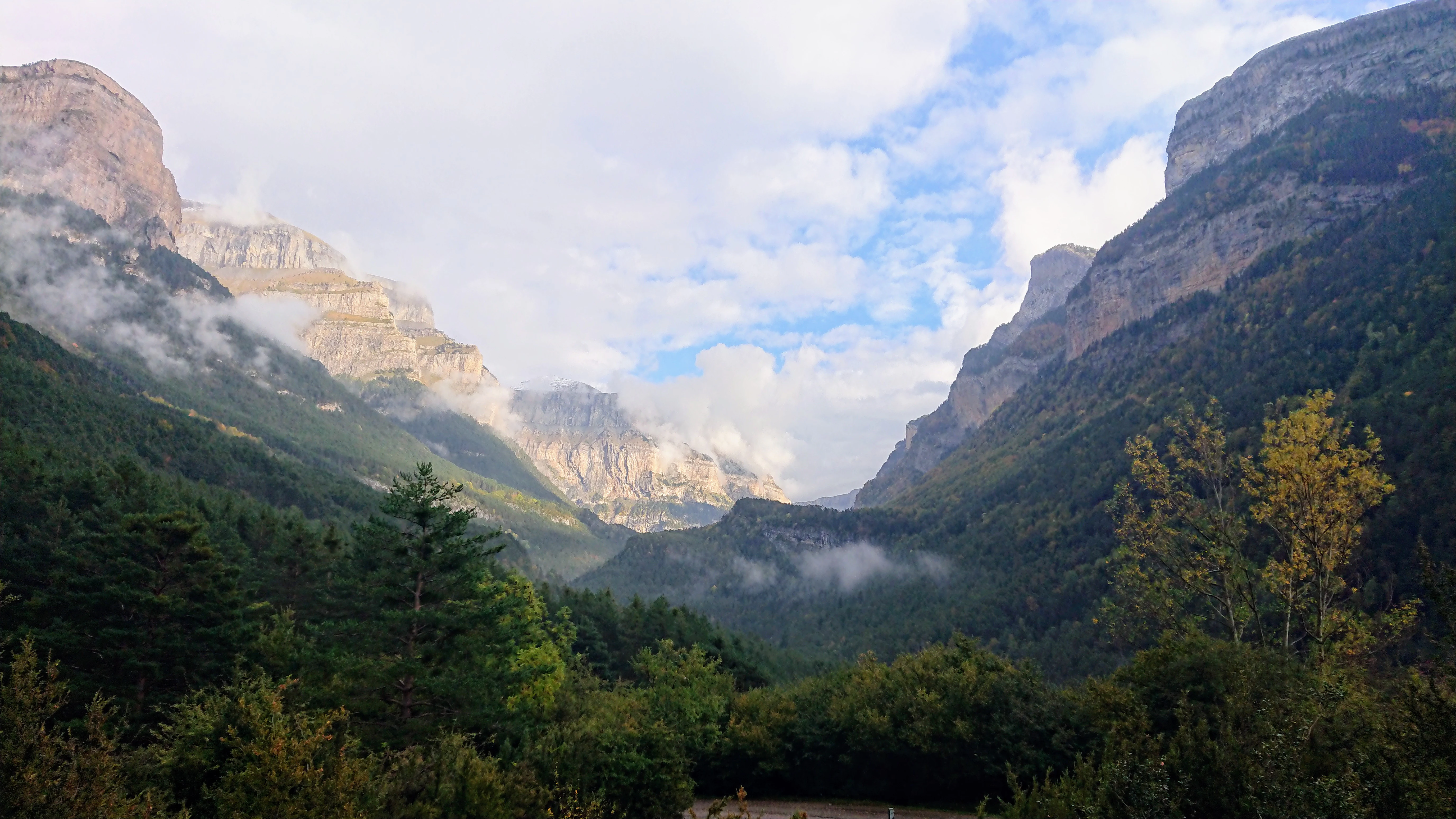 Landscape view of Ordesa Valley with towering mountain peaks, forested slopes, and misty valleys under a partly cloudy sky.