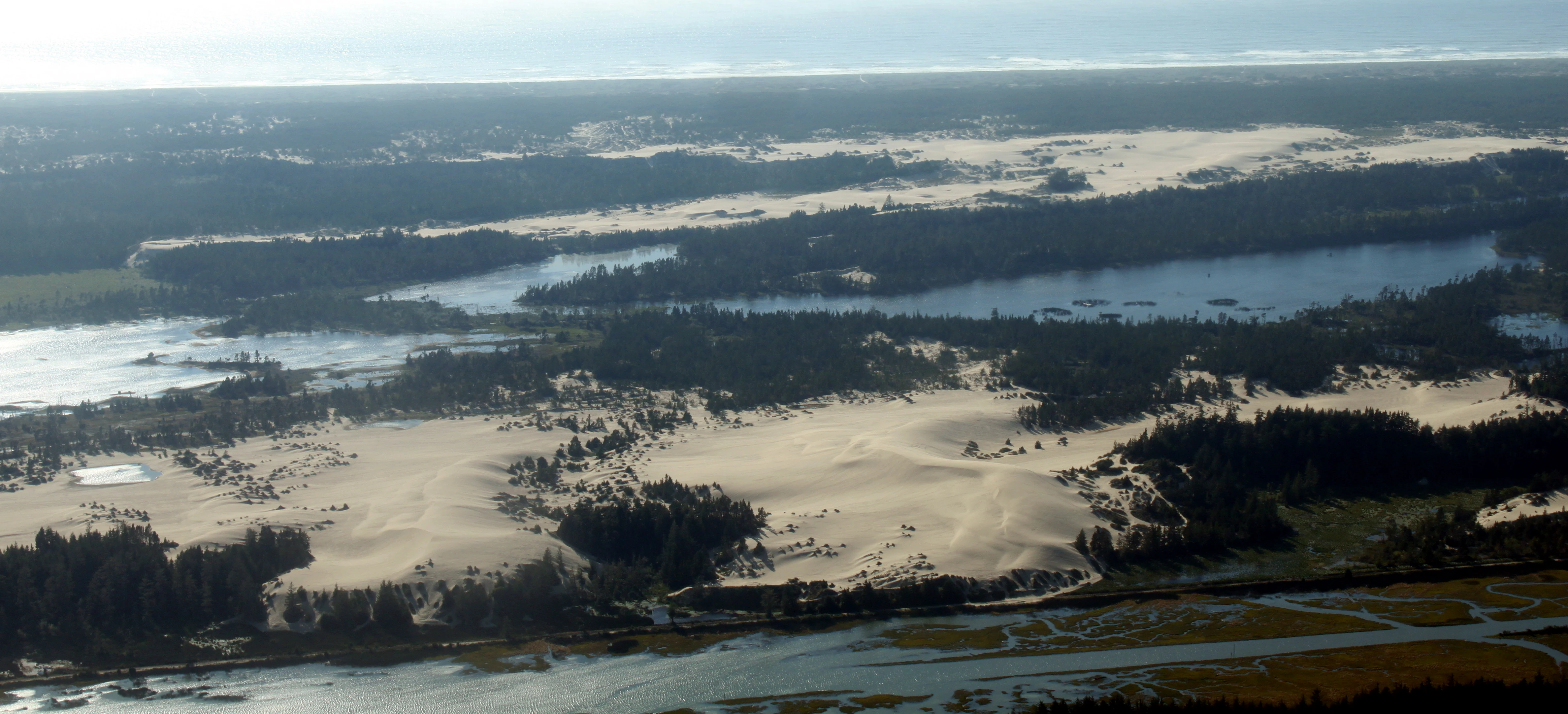 Aerial view looking over a large landscape featuring vast patches of light-colored sand dunes separated by dark green forests and coastal waterways.
