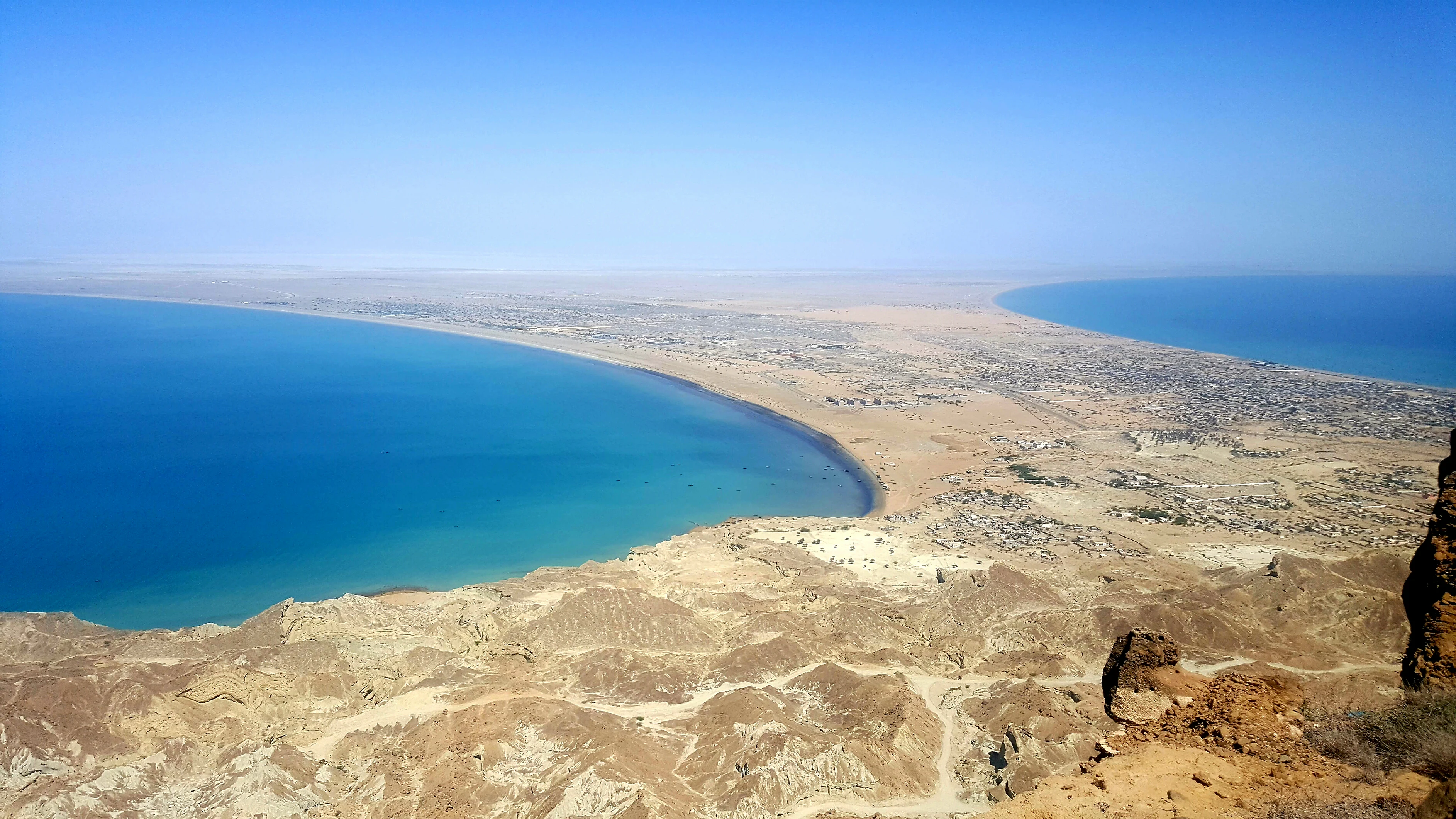 A wide aerial view looking down from a high rocky ridge over a coastal land bridge flanked by two crescent-shaped blue bays and arid terrain.