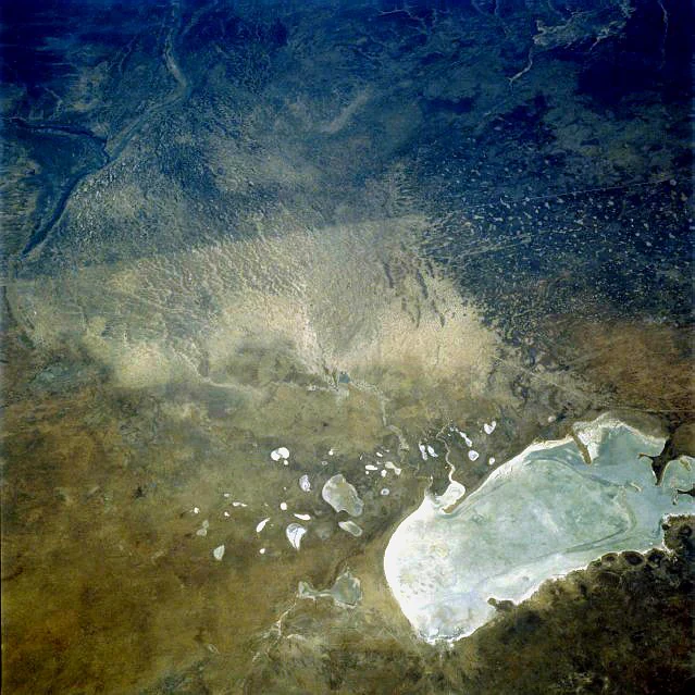 An aerial view showing a vast, bright white salt pan contrasted against dark, arid surrounding terrain with scattered smaller pools and drainage patterns.