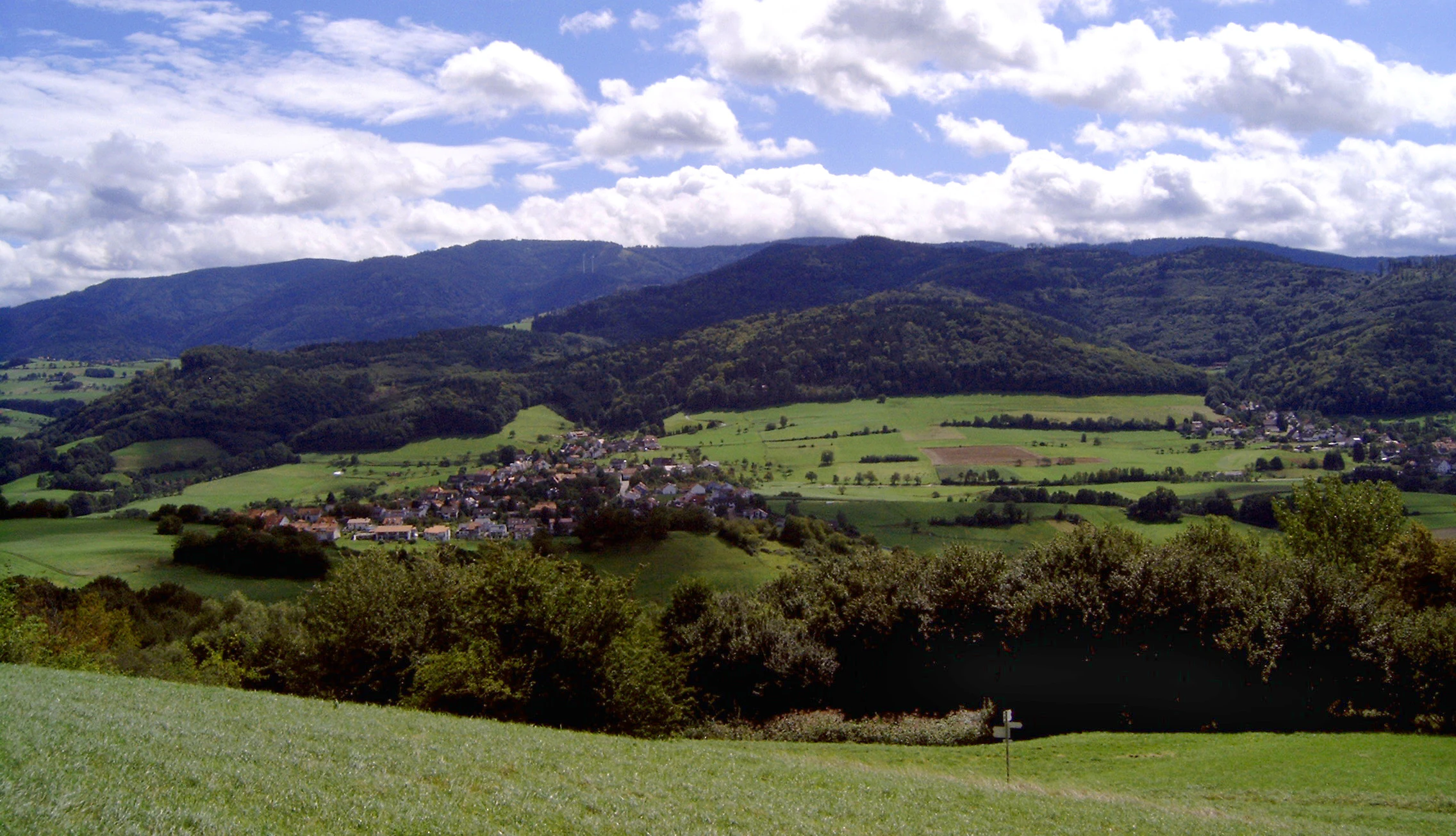 A wide landscape photograph of a green valley with a small village, surrounded by forests and rolling hills, with a large forested mountain in the background under a blue sky with white clouds.