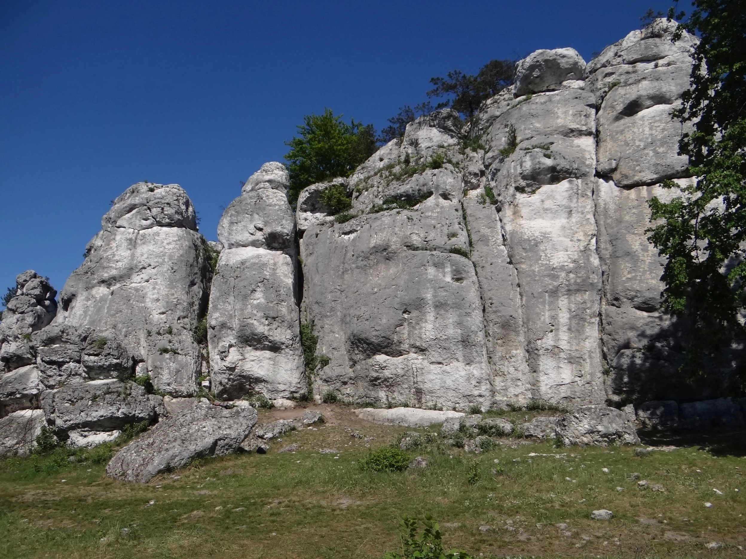 A large, light-colored limestone rock formation with vertical fissures rises above a grassy slope under a clear blue sky.