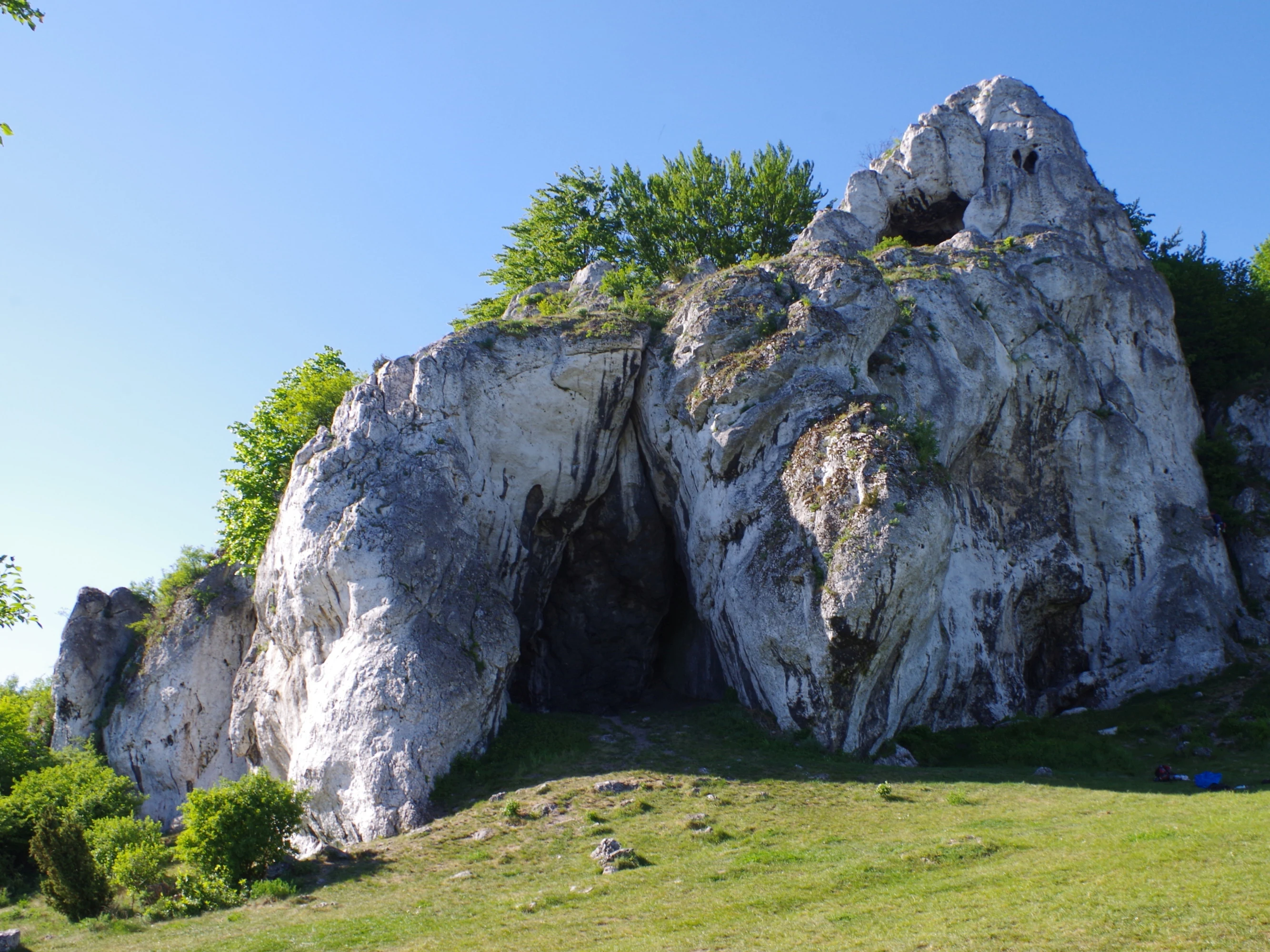 A large, pale limestone rock formation with a natural cave entrance set against a bright blue sky, surrounded by green trees and grass.