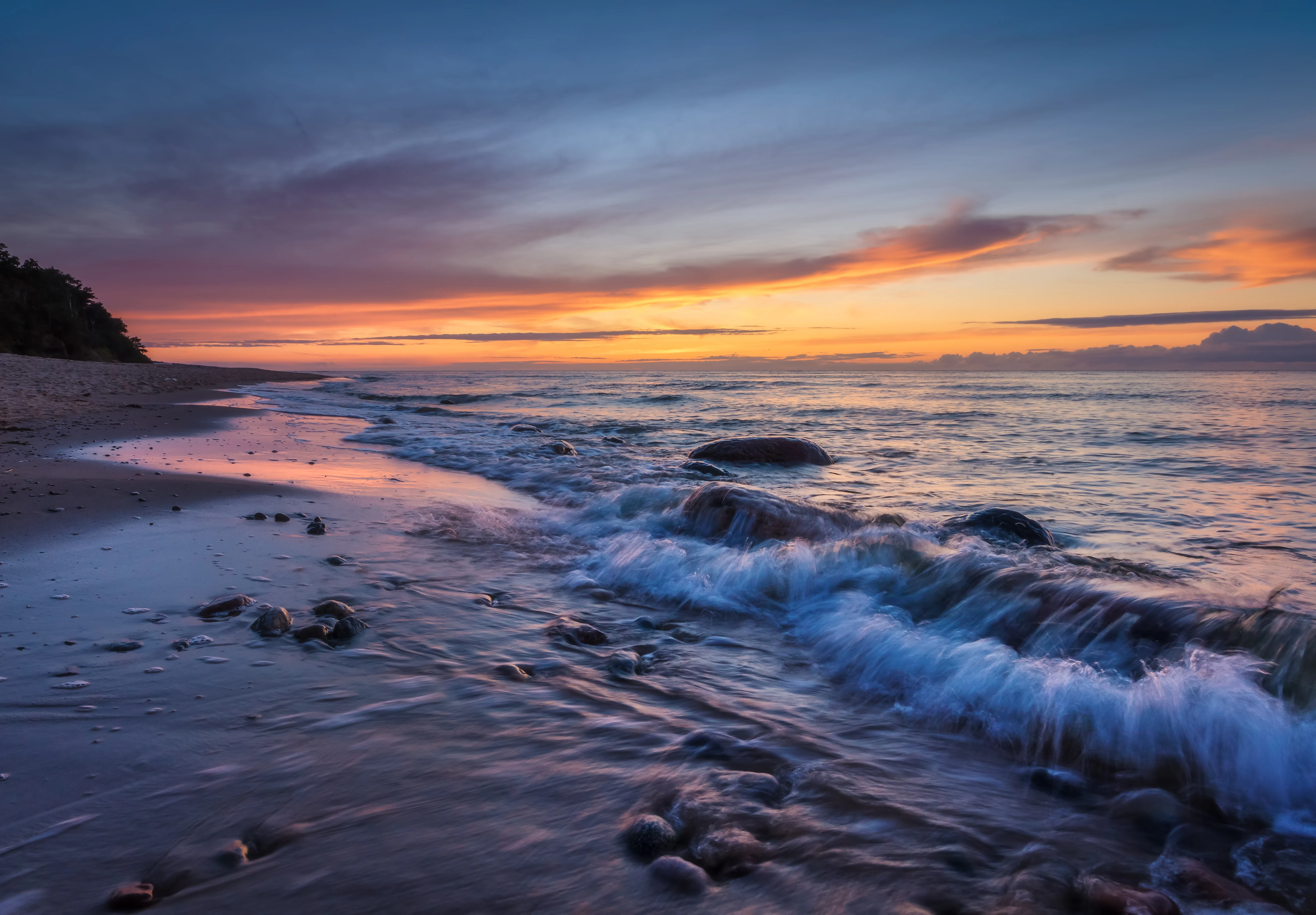 A wide angle view of a sandy beach meeting the Baltic Sea at sunset, with waves washing over stones in the foreground.