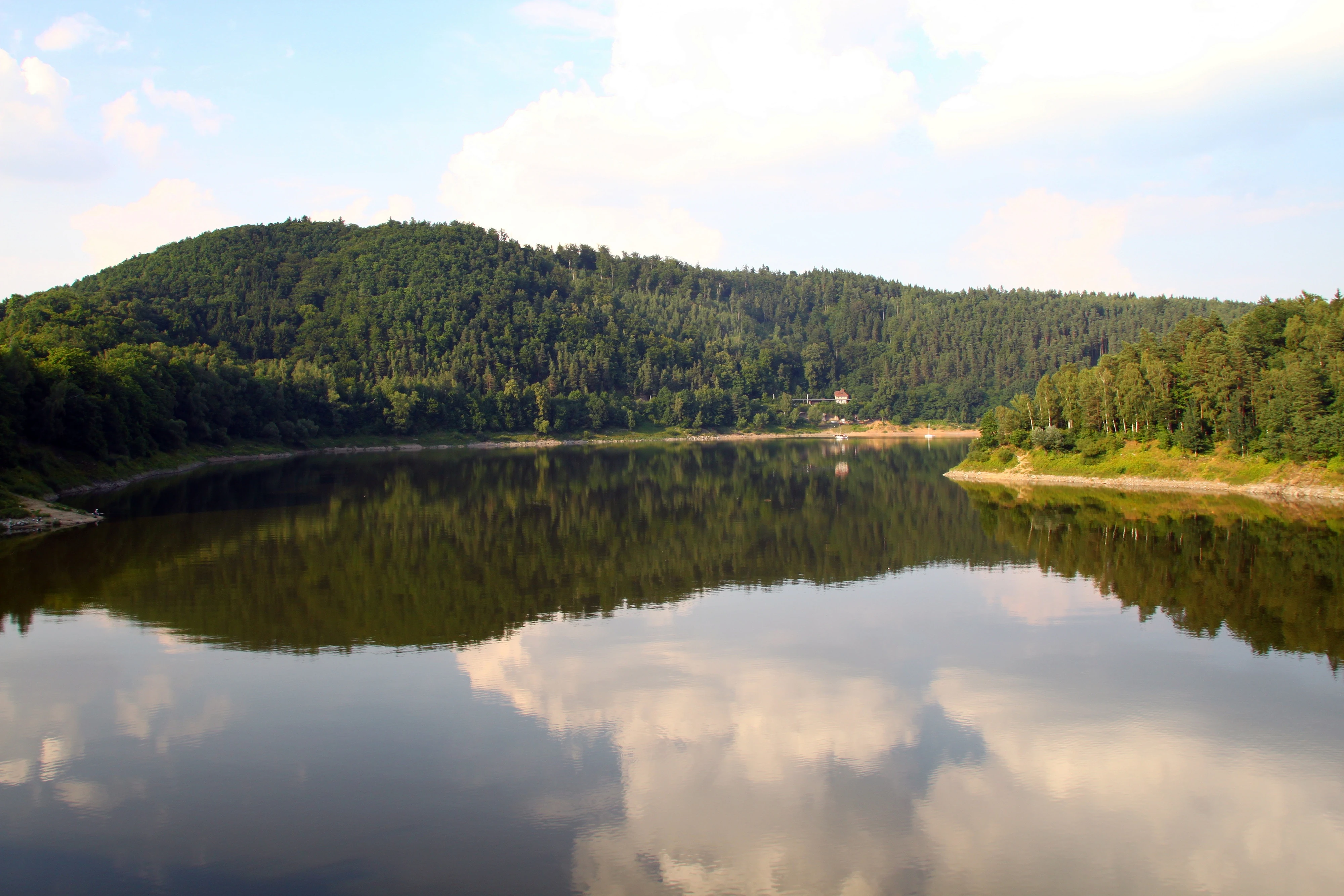 A wide, calm river or reservoir reflecting a bright, cloudy sky and dense, green forested hills on its banks.