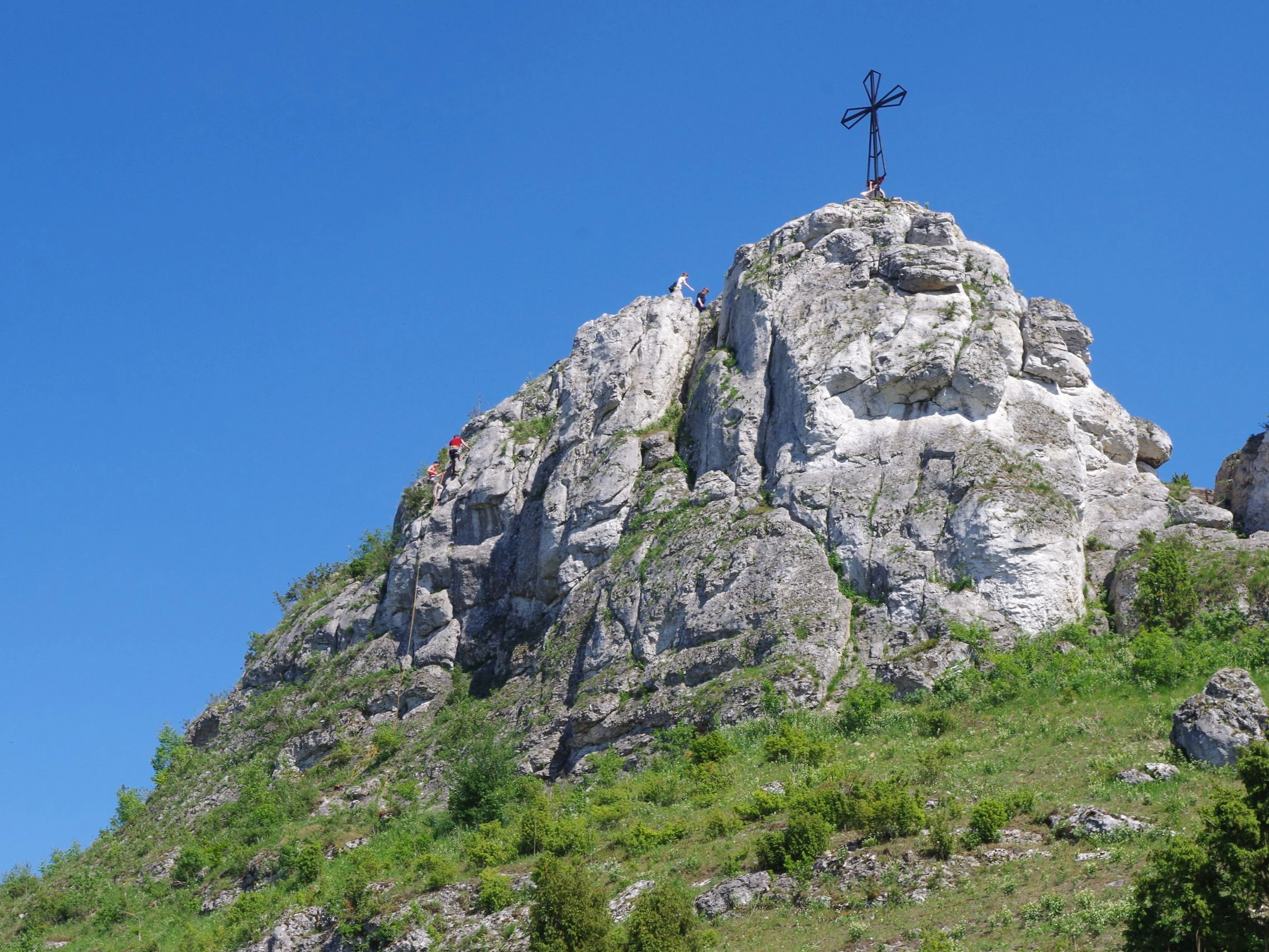 A steep, light-colored limestone rock formation topped with a metal cross under a clear blue sky, with greenery at its base.