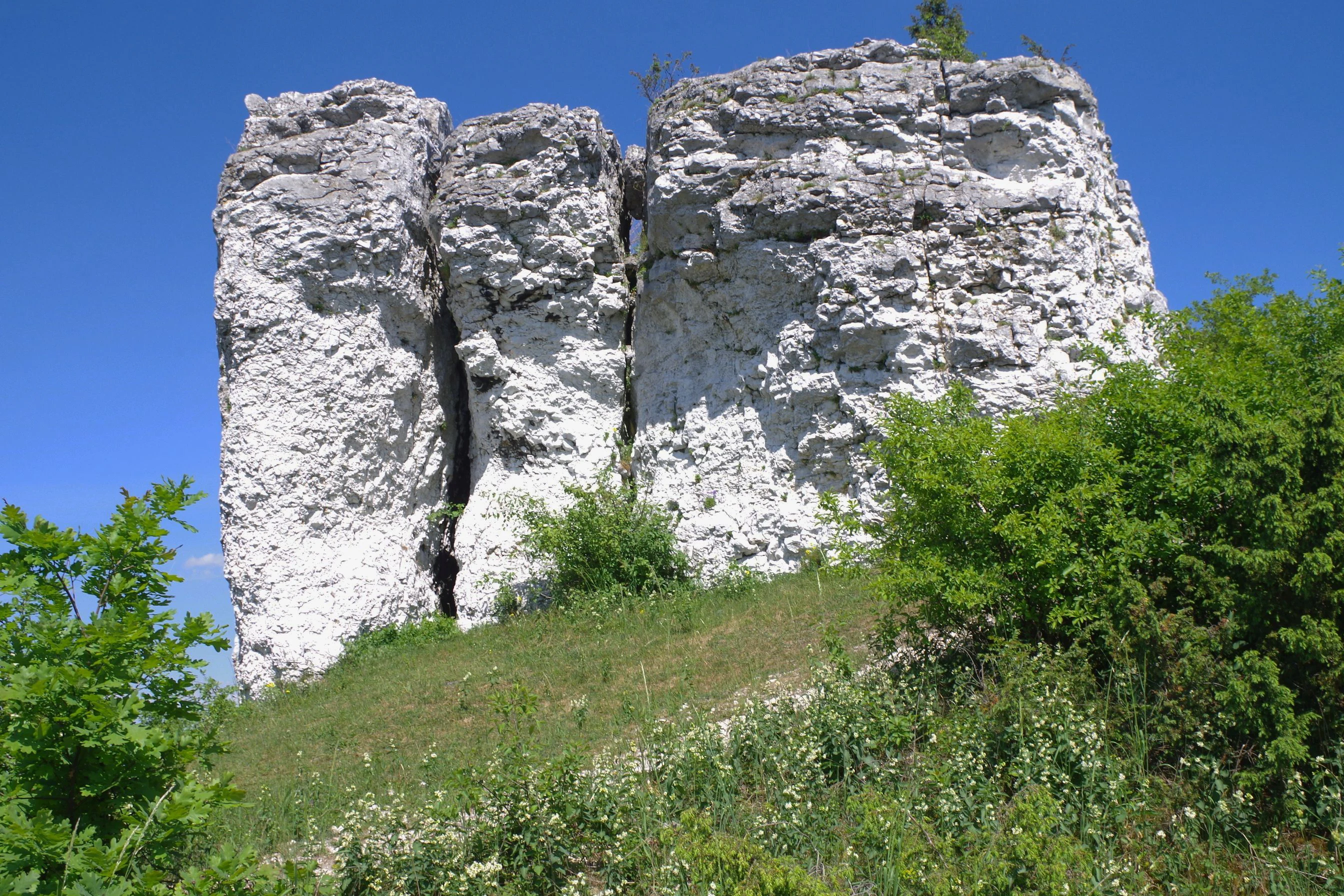 A weathered white limestone rock formation rising from a grassy, sunlit hillside under a clear blue sky.