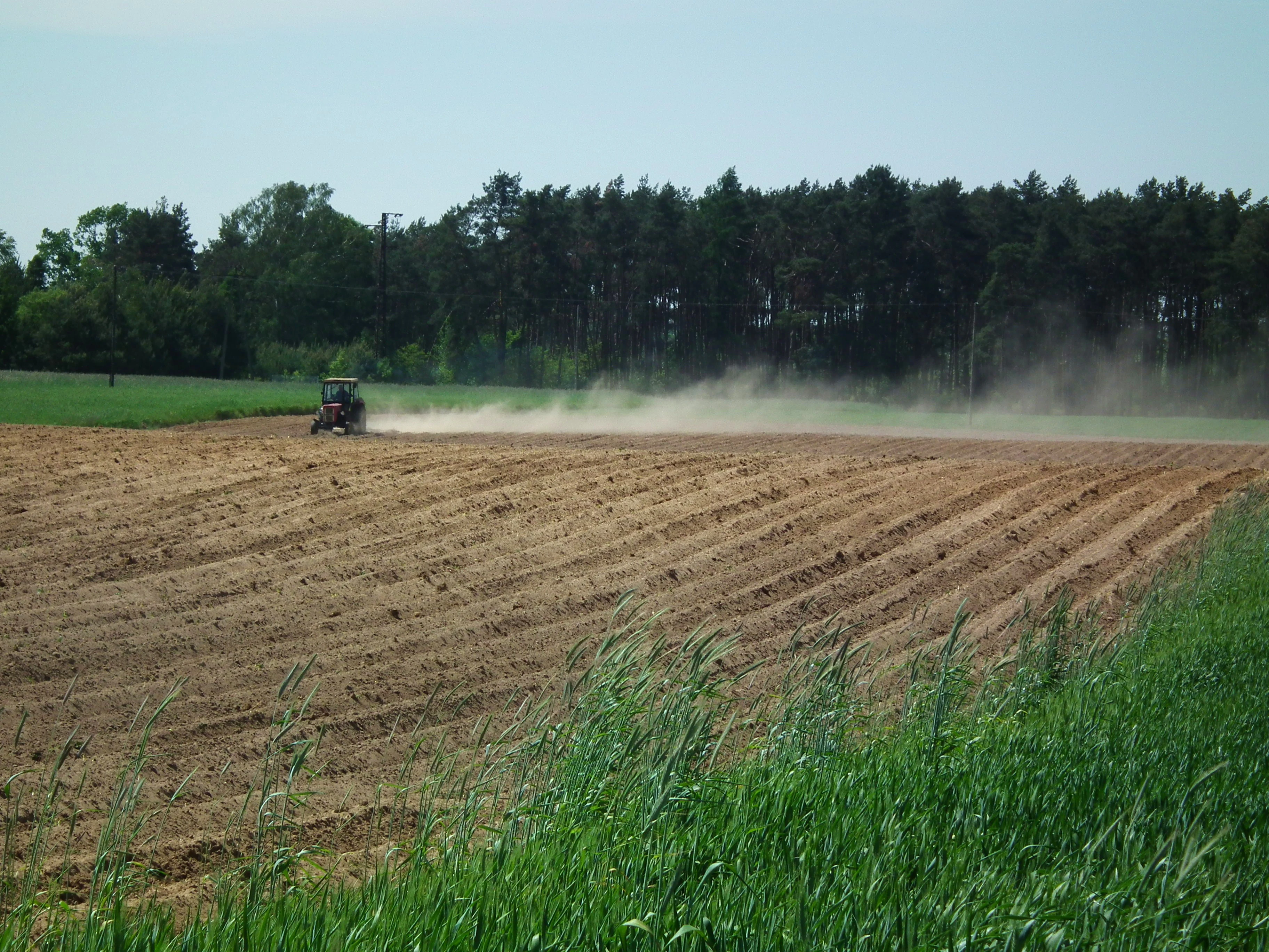 A tractor plowing a field, creating a cloud of dust, with a line of trees in the background.