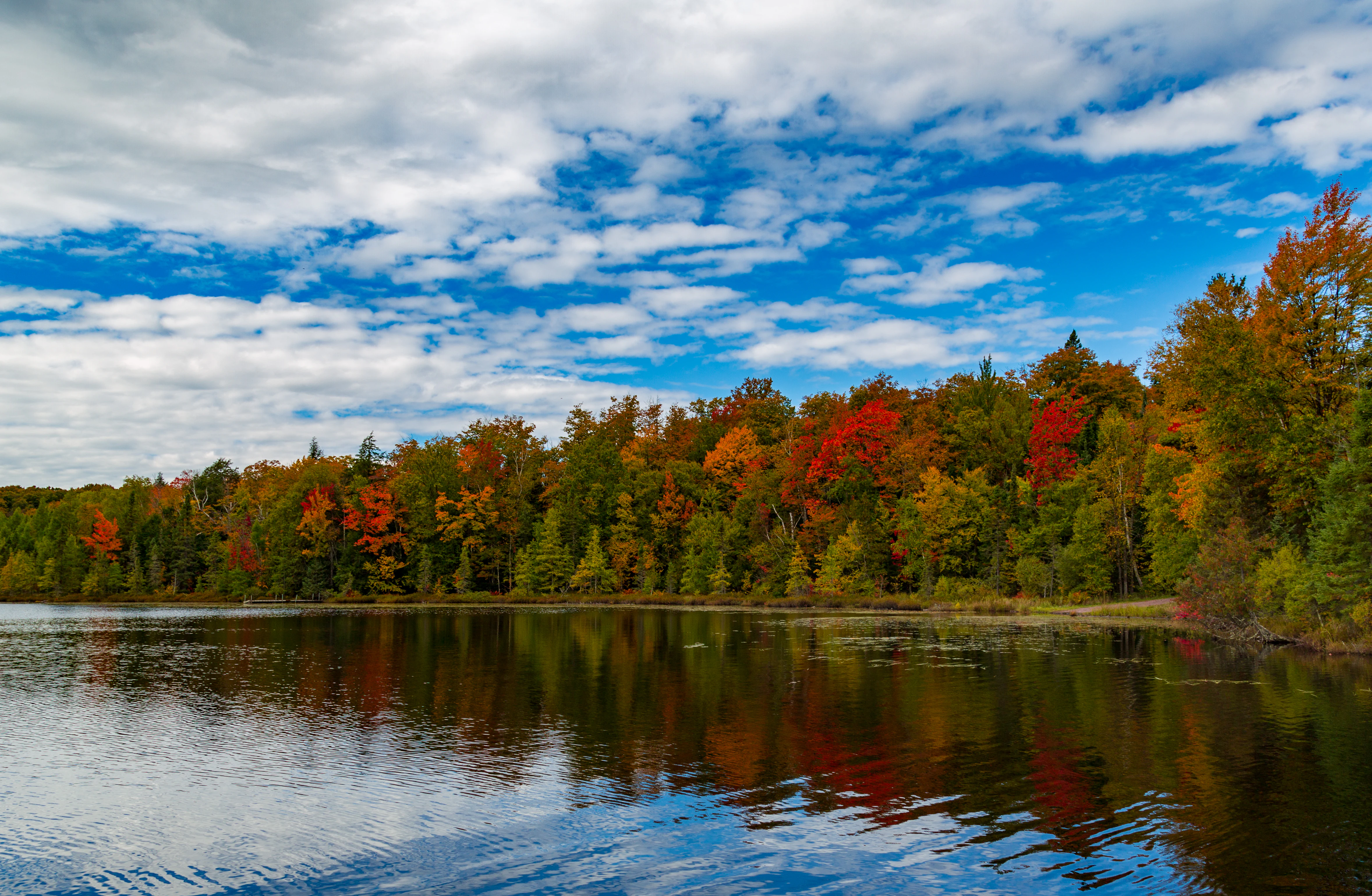 A peaceful lake in autumn with vibrant red, orange, and yellow fall foliage on the forested shoreline reflecting in the calm water under a partly cloudy blue sky.