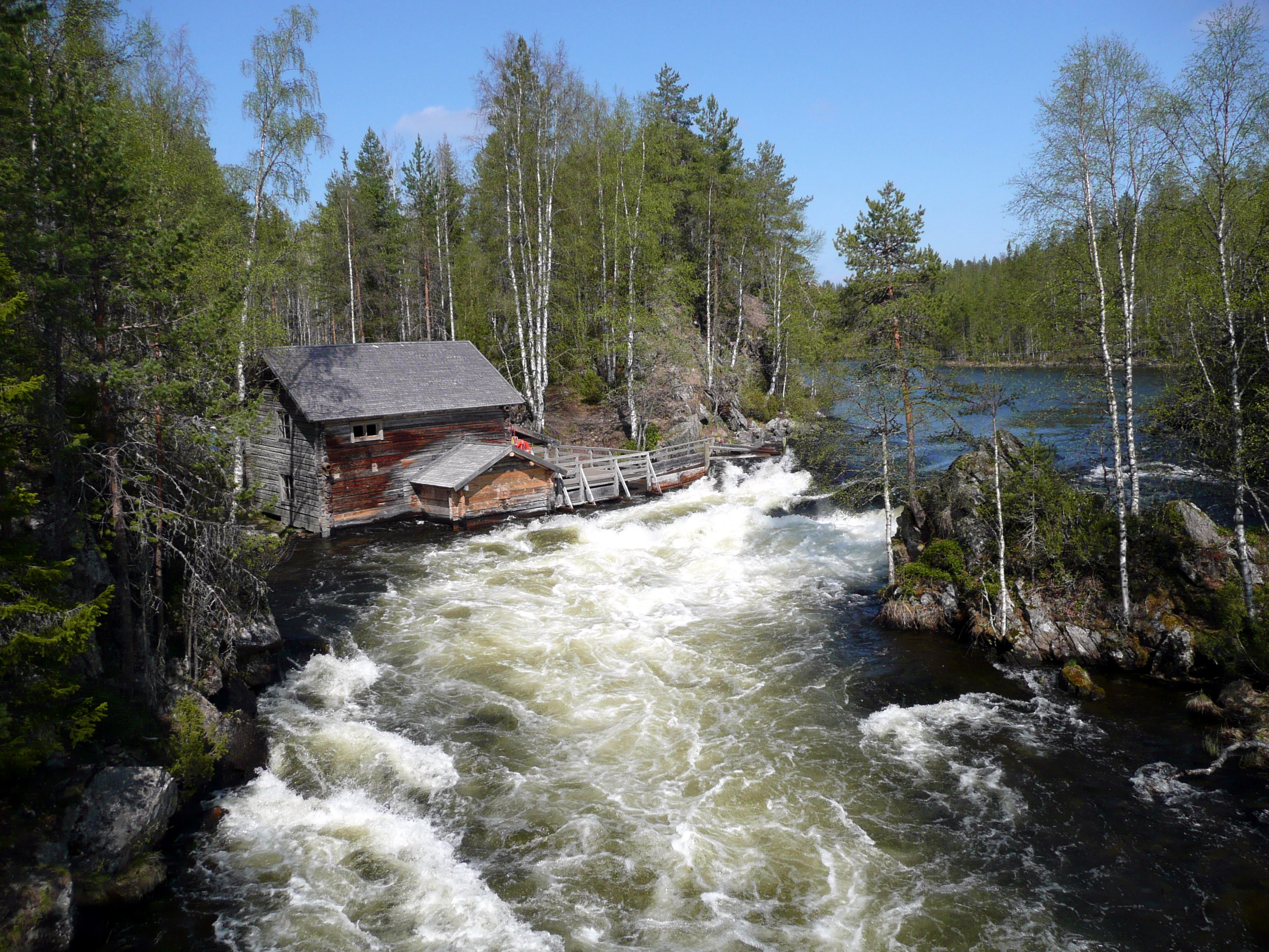 Rapid river flow with white water, wooden building with gray roof beside the river, surrounded by forest trees under clear blue sky