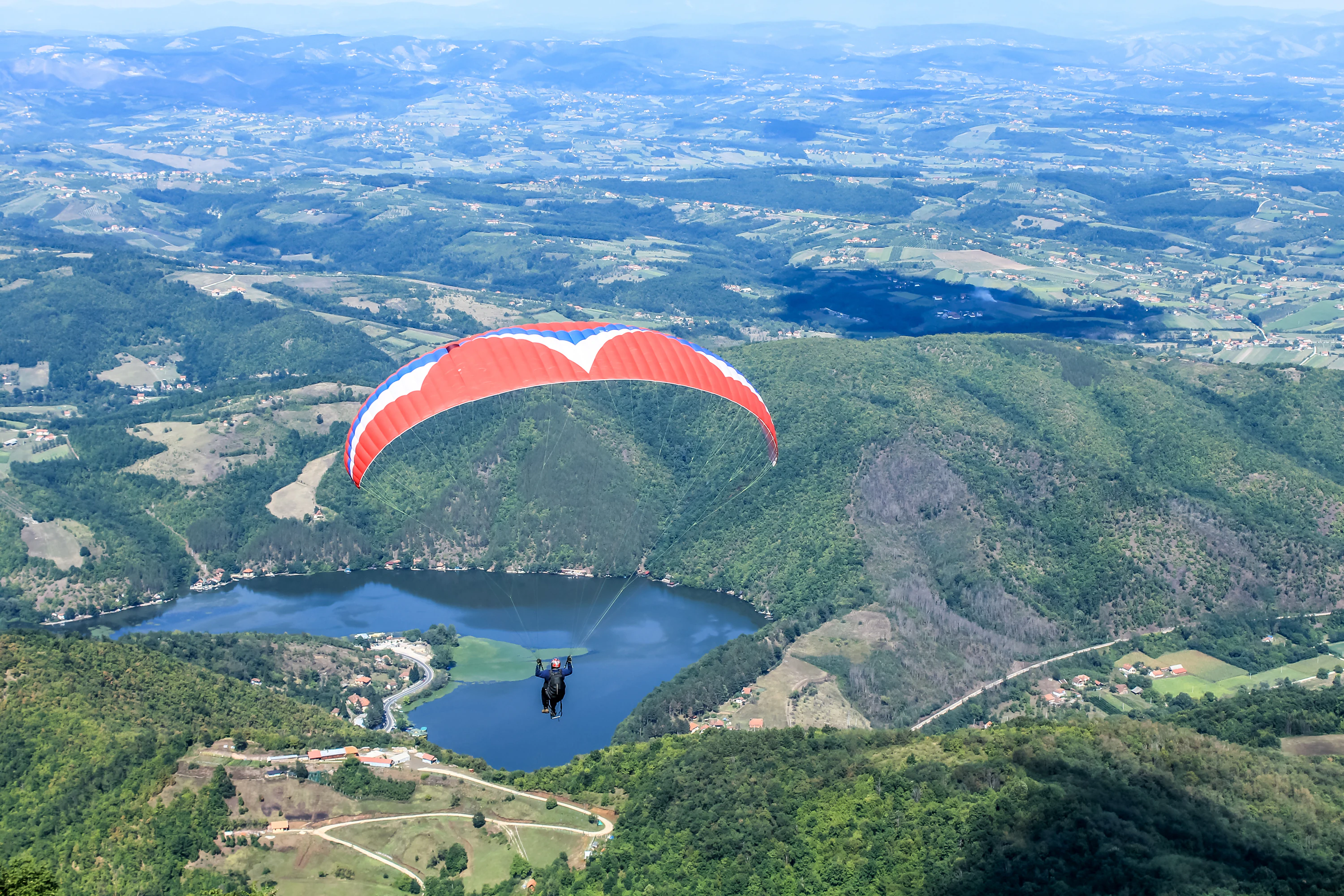 A red and white paraglider flying over a winding river valley surrounded by green hills and mountains