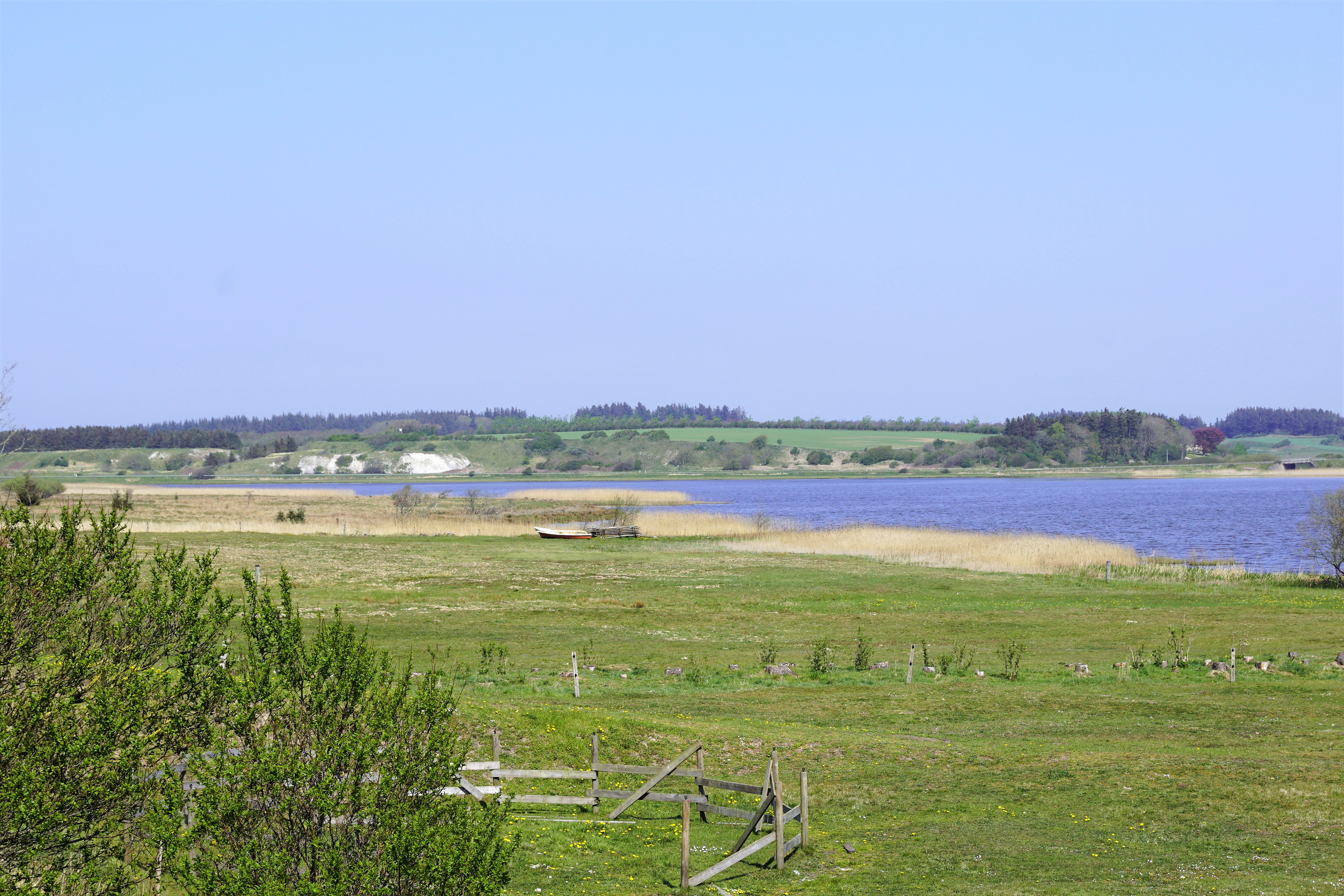 A wide landscape view of a calm blue lake, Ove Sø, surrounded by open grassy meadows and distant trees under a clear blue sky.