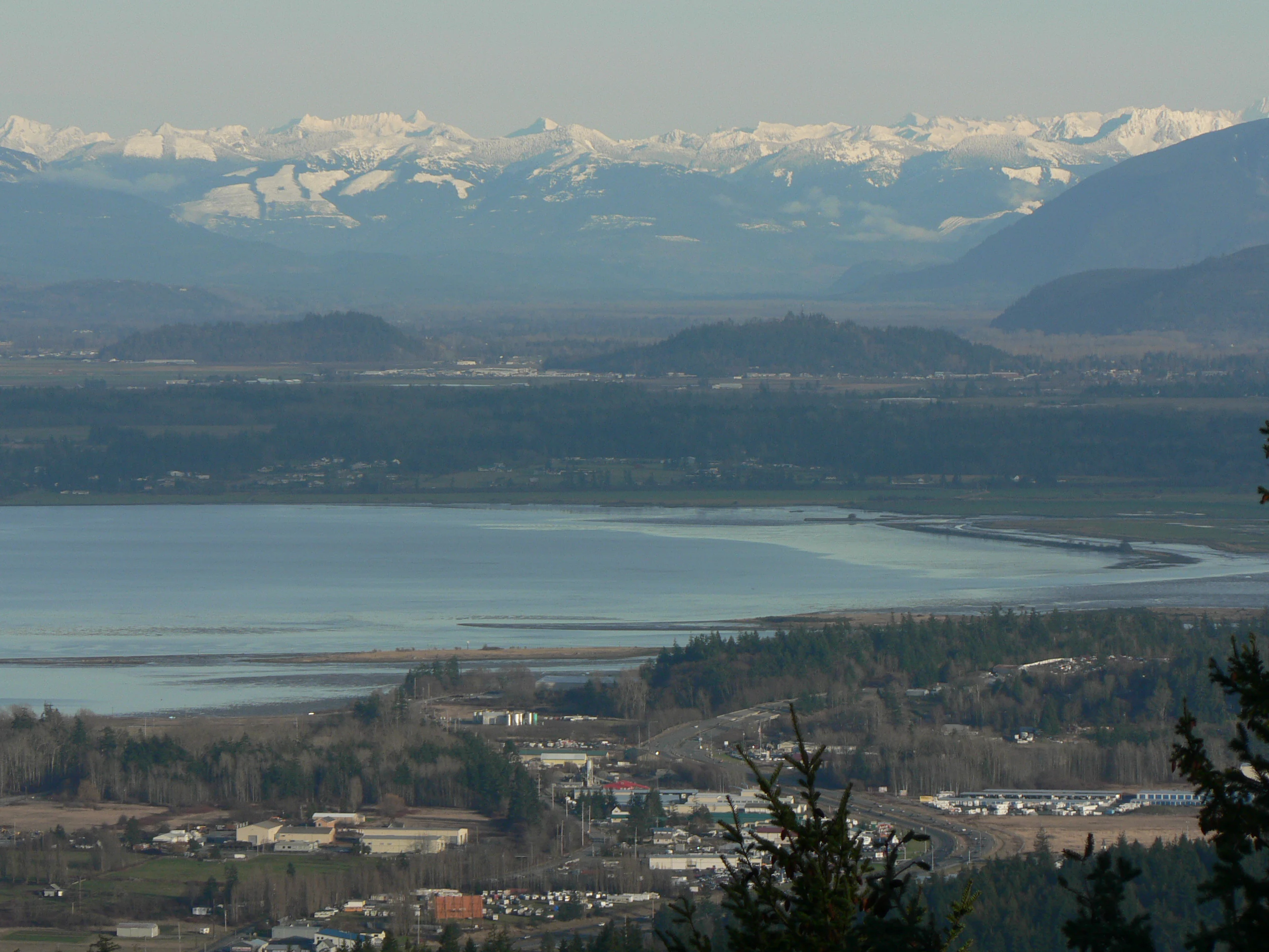 A wide aerial view showing Padilla Bay in the middle distance, with a flat valley floor and snow-capped mountain range spanning the horizon.
