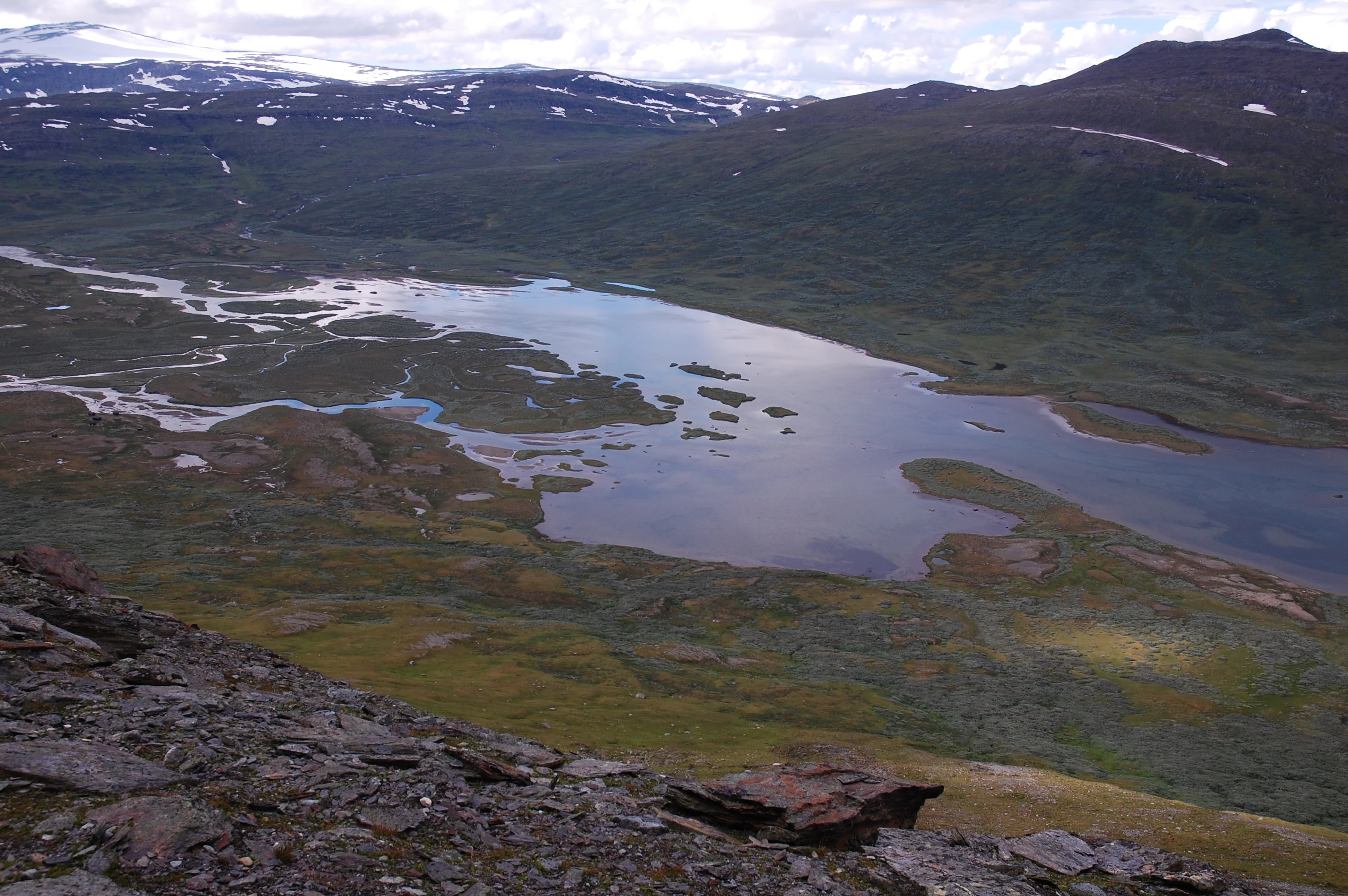 Wide view of Kapasluoppal Lake surrounded by mountainous terrain with patches of snow, green and brown vegetation, and rocky foreground
