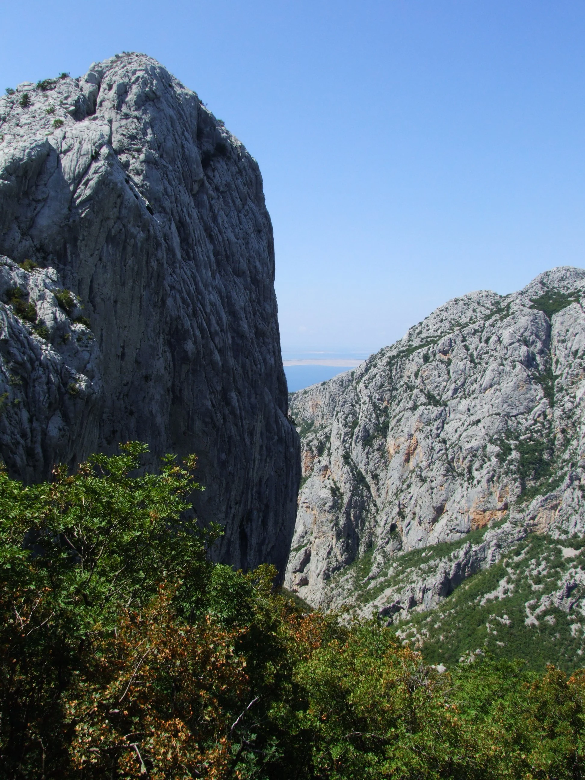 A rocky mountain peak with steep cliffs, green vegetation in the foreground, and a view of the sea under a clear blue sky