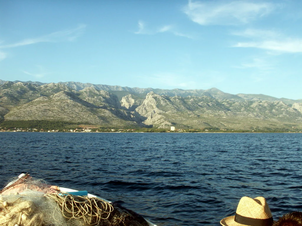 Blue sea with rocky mountains in the background, viewed from a boat. Fishing nets and a straw hat are visible in the foreground.