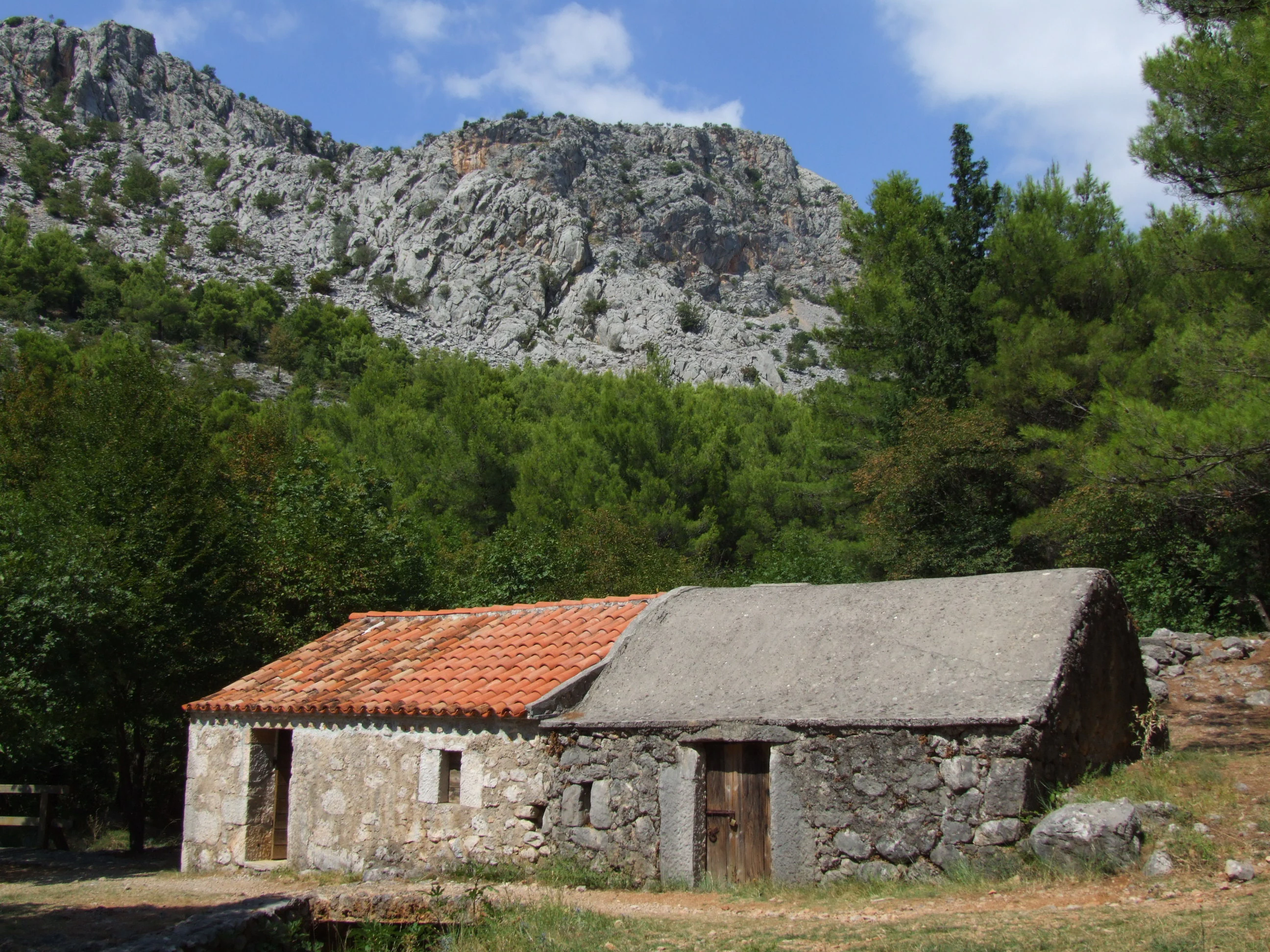 Stone building with red-tiled roof and adjacent stone structure with gray roof, set against rocky mountains and green trees