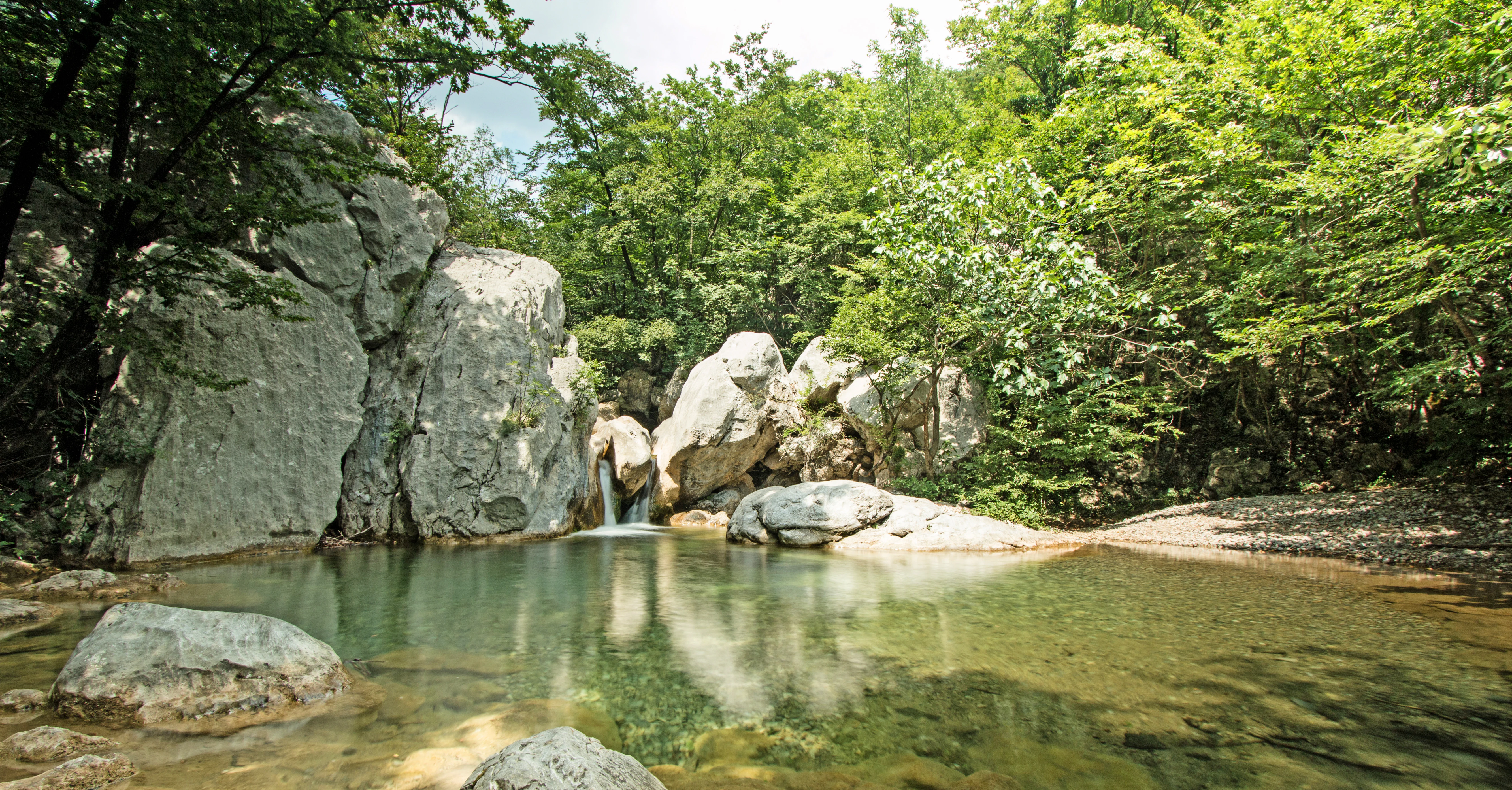 Clear water pool with small waterfall between large rocks surrounded by dense green forest