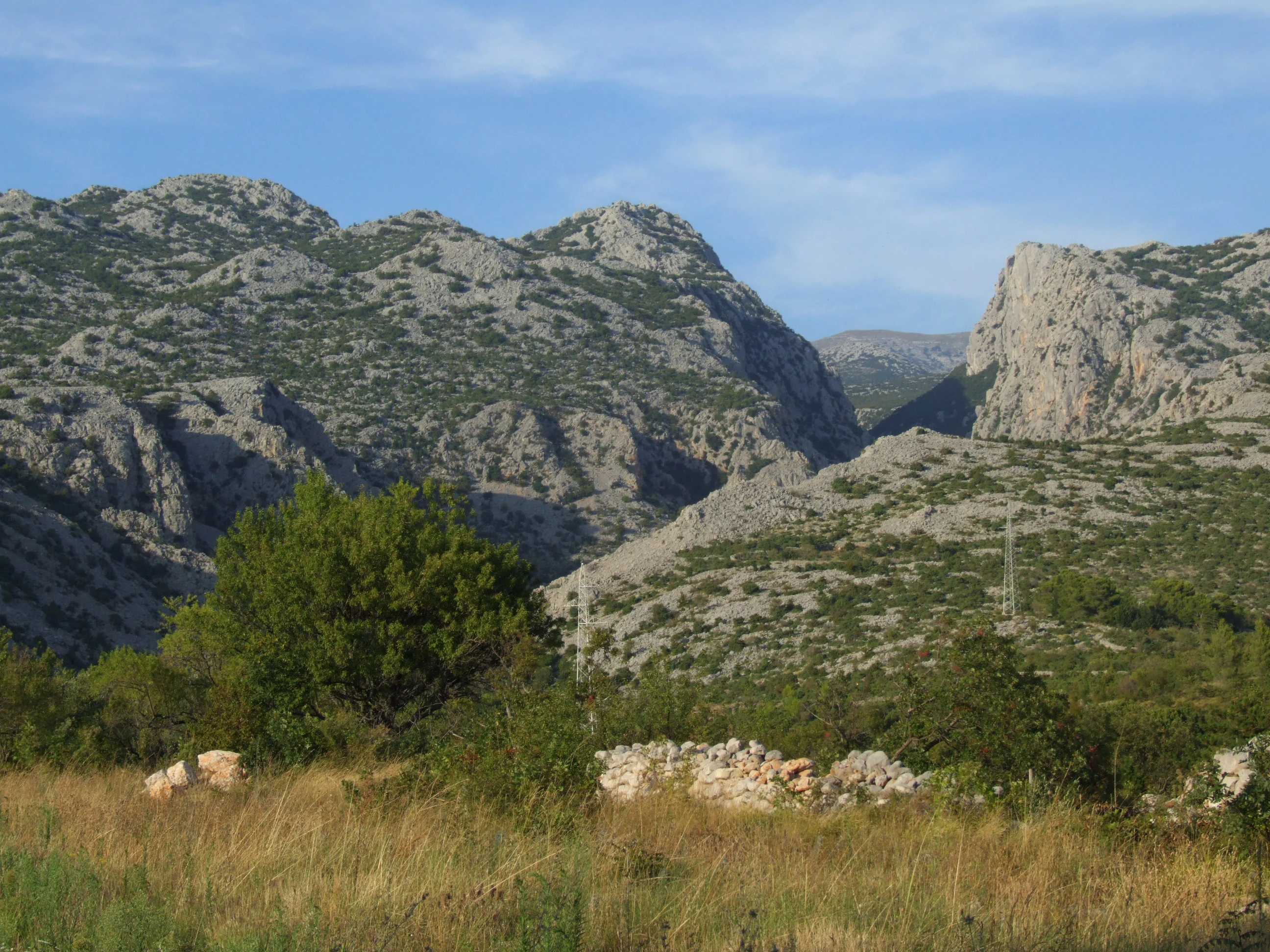 Rocky mountain peaks with sparse vegetation, stone wall, and grassy field in foreground under clear sky