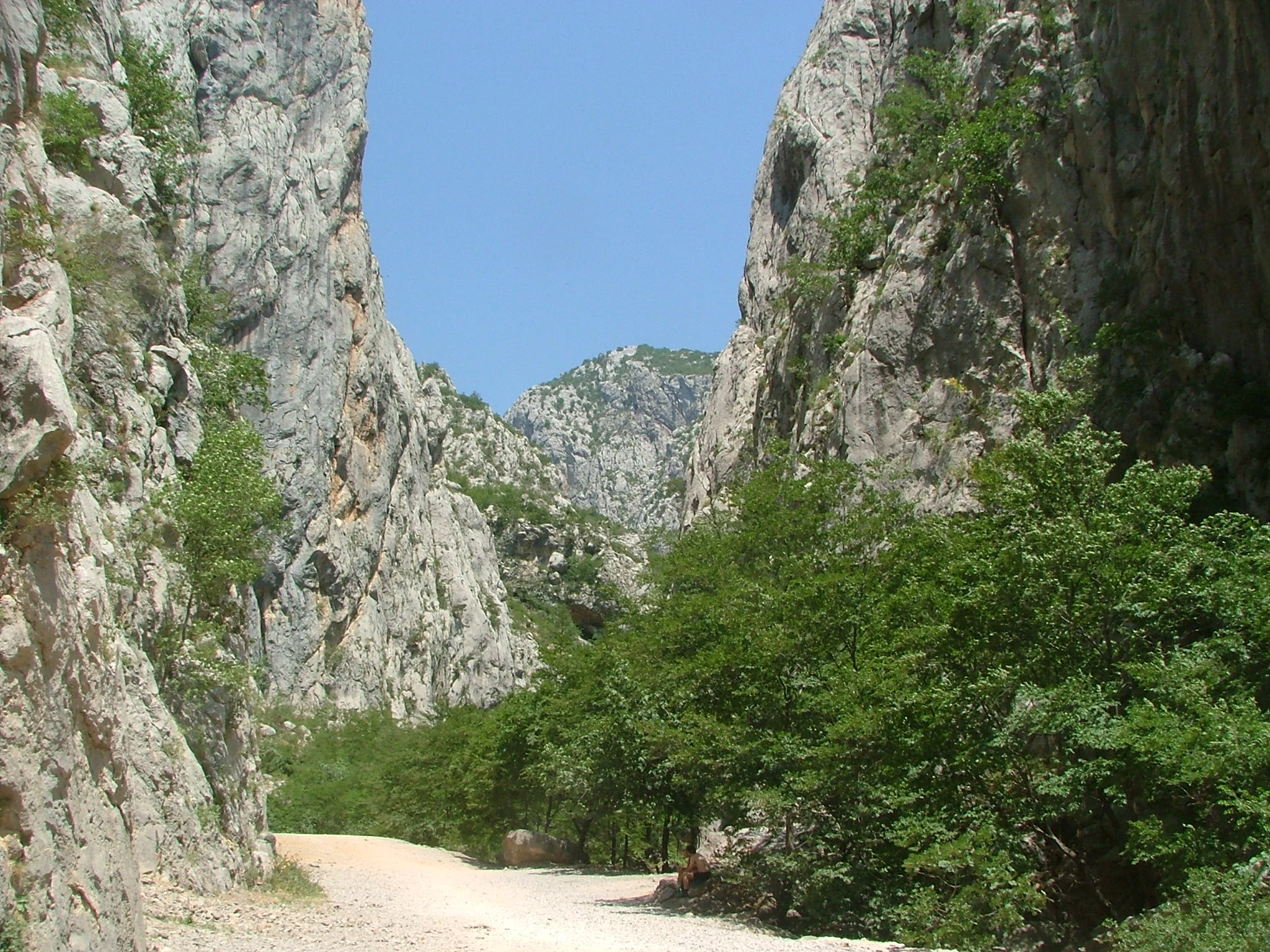 Steep rocky cliffs on both sides of a valley with green vegetation and a dirt path under a clear blue sky