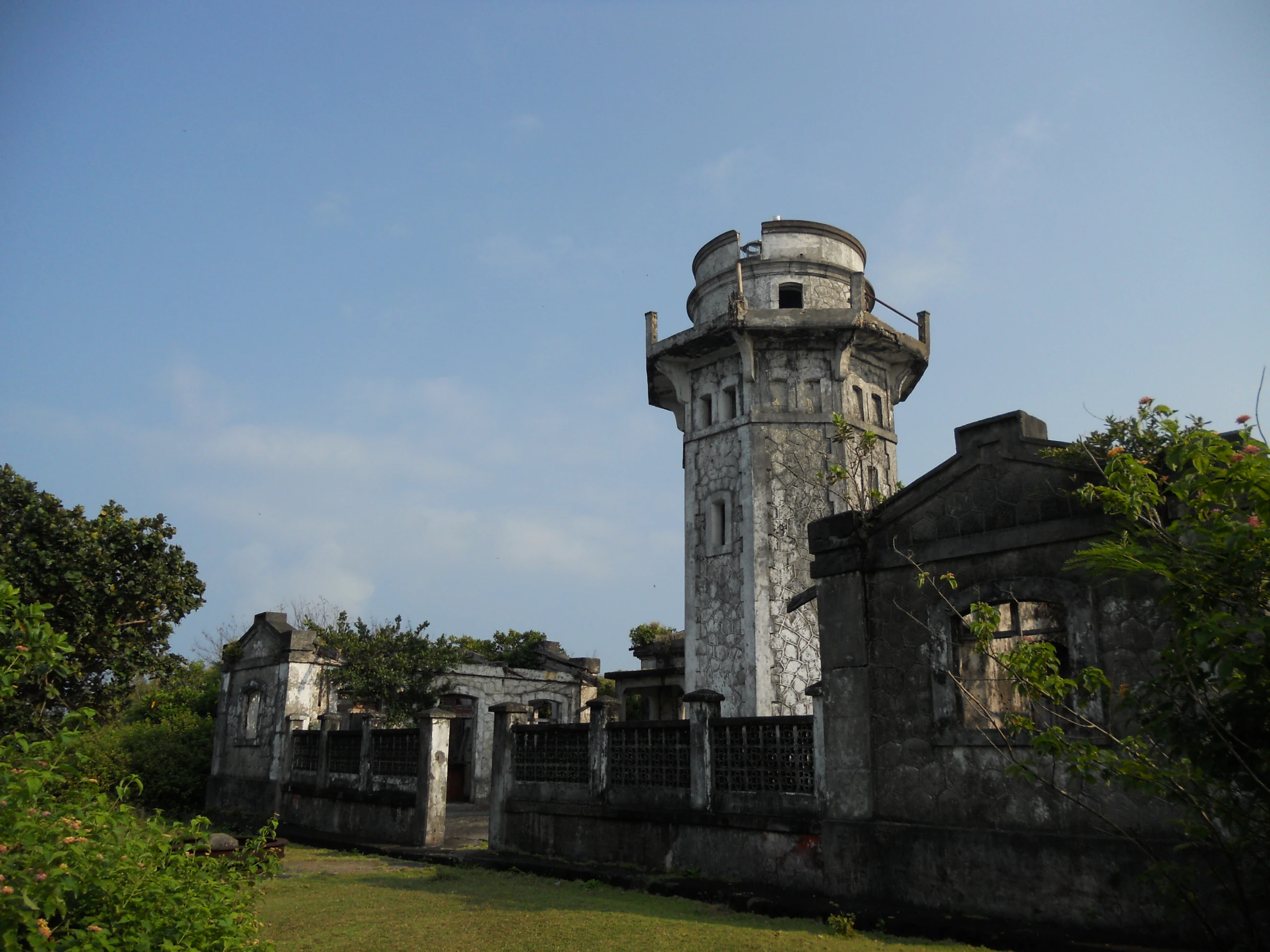 A view of the historic, weathered stone ruins of the Cape Engano Lighthouse tower and surrounding structures against a clear blue sky.
