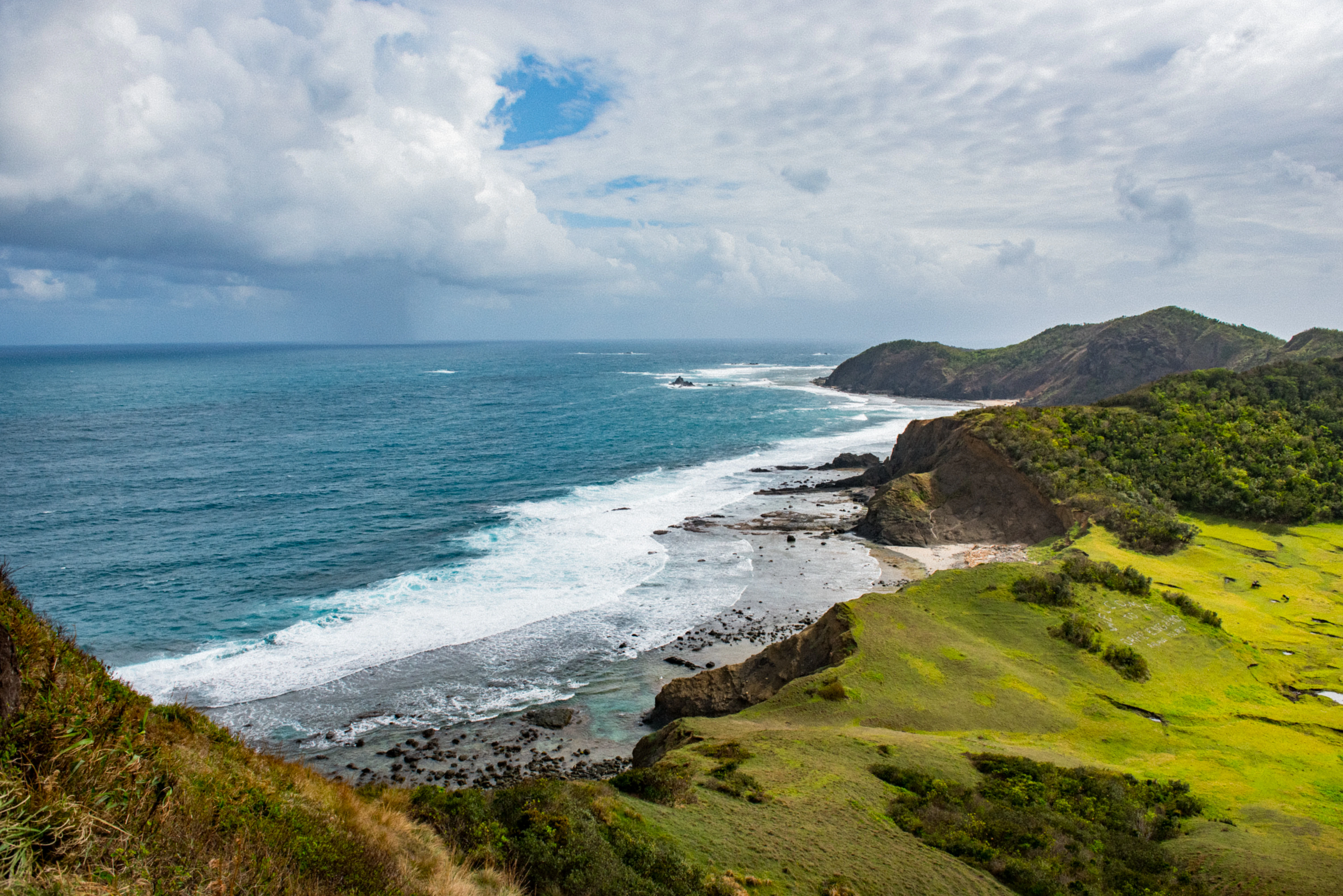 An expansive coastal landscape overlooking the ocean, featuring bright green grassy hills in the foreground and rocky cliffs leading to a sandy beach with crashing waves under a cloudy sky.
