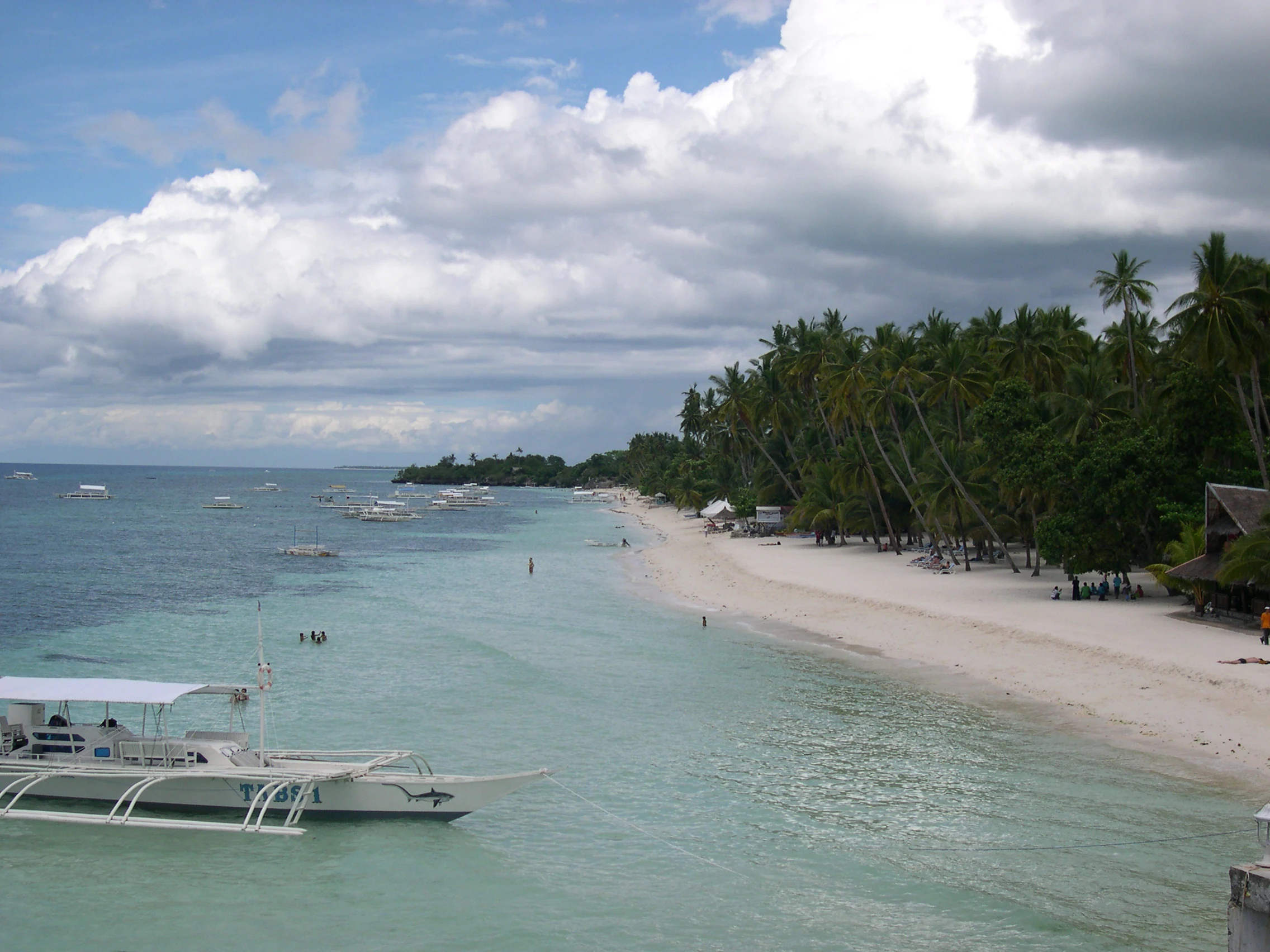 A wide-angle scenic view of a tropical white sand beach with palm trees along the shore, clear turquoise shallow water, and several traditional Filipino outrigger boats floating in the water.