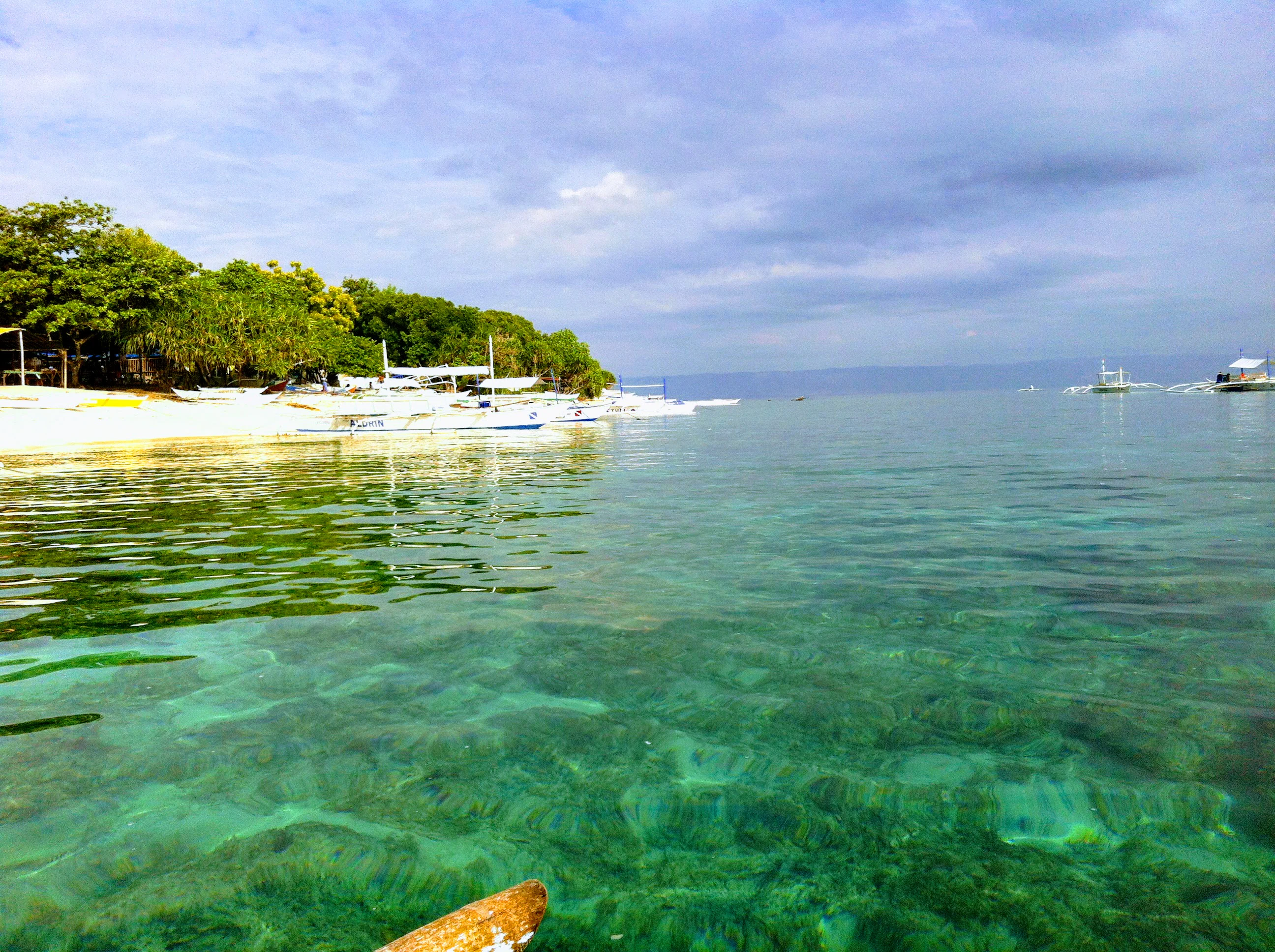 A view from the water looking toward a coastline with shallow coral reefs visible beneath the surface and outrigger boats moored near a sandy beach.