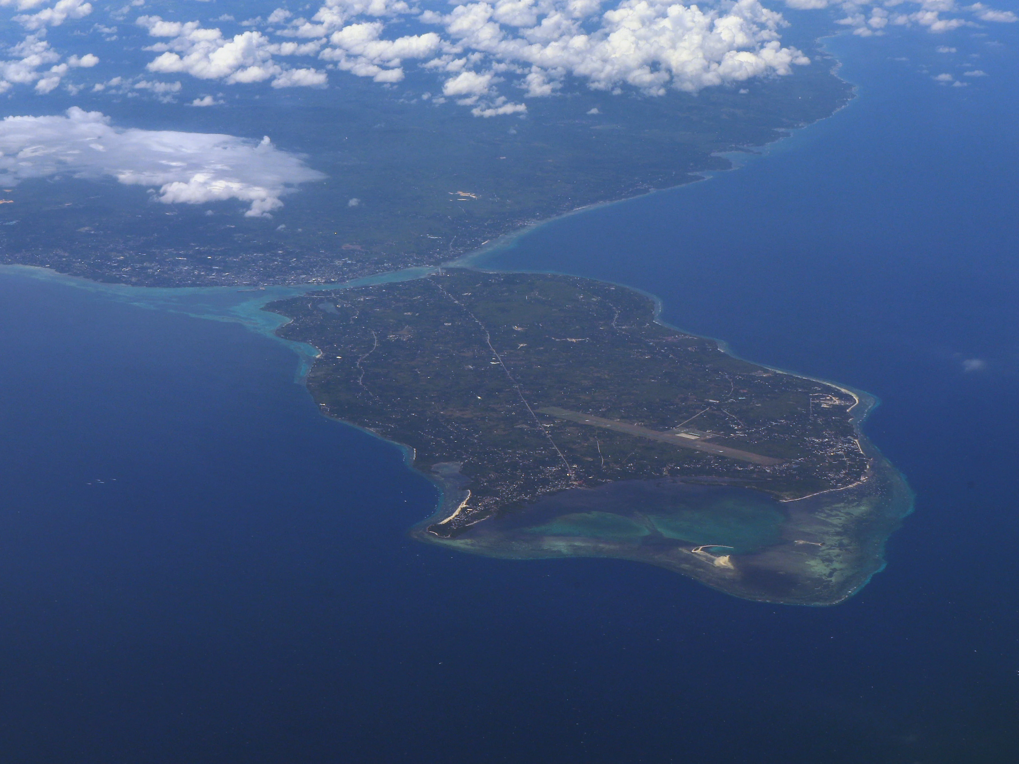 An aerial view of an island surrounded by deep blue ocean water, with a clear separation between the island landmass, shallow turquoise coral reefs, and the deep sea.