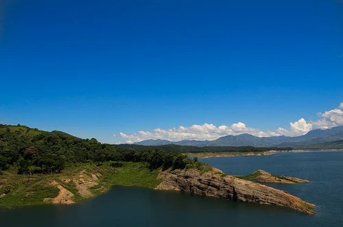 A wide, elevated view of a large reservoir with calm blue water, rocky shoreline, and distant mountains under a clear blue sky.
