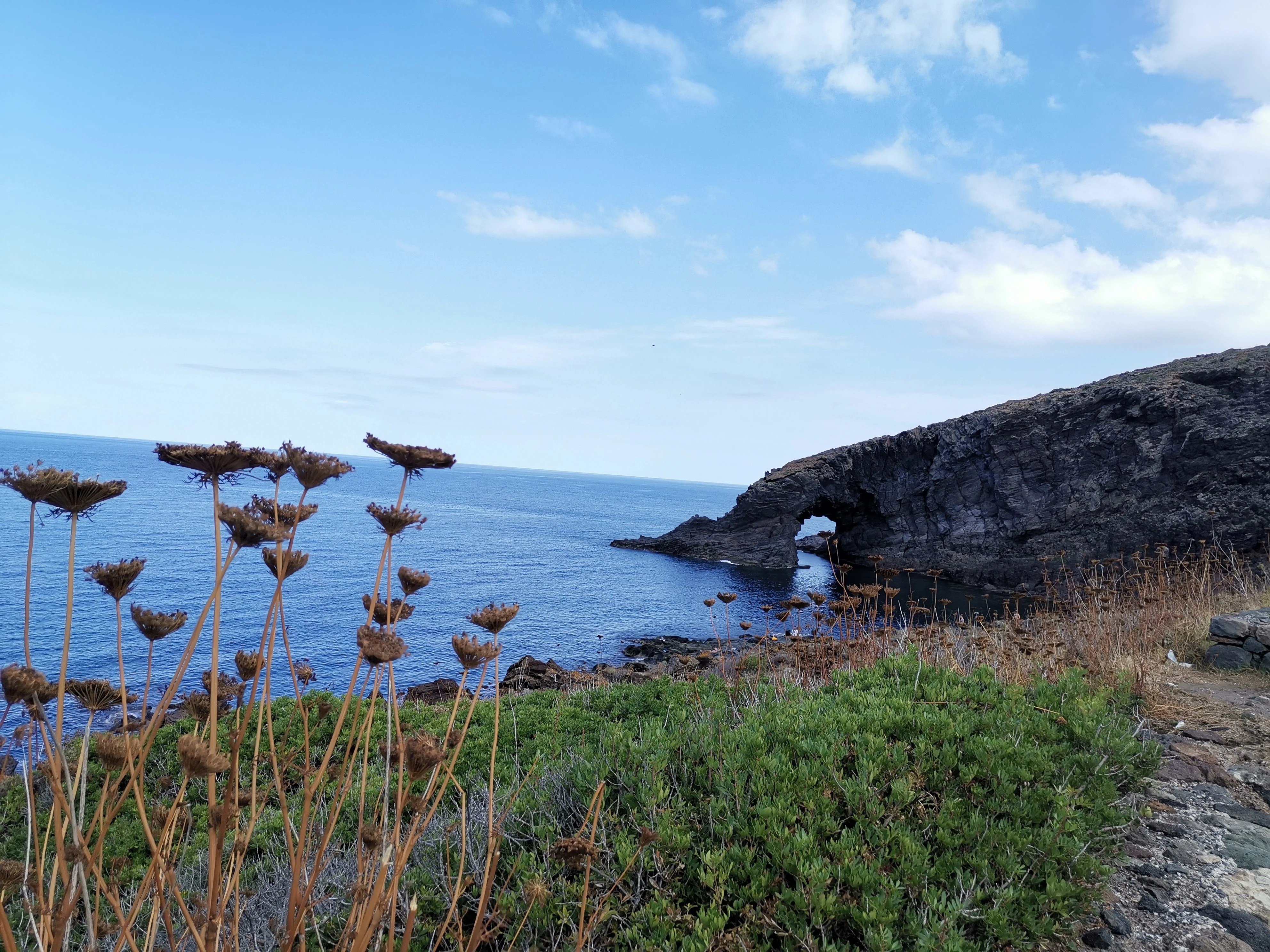 Natural rock arch on coastal cliffs overlooking blue sea with dry vegetation and clear sky