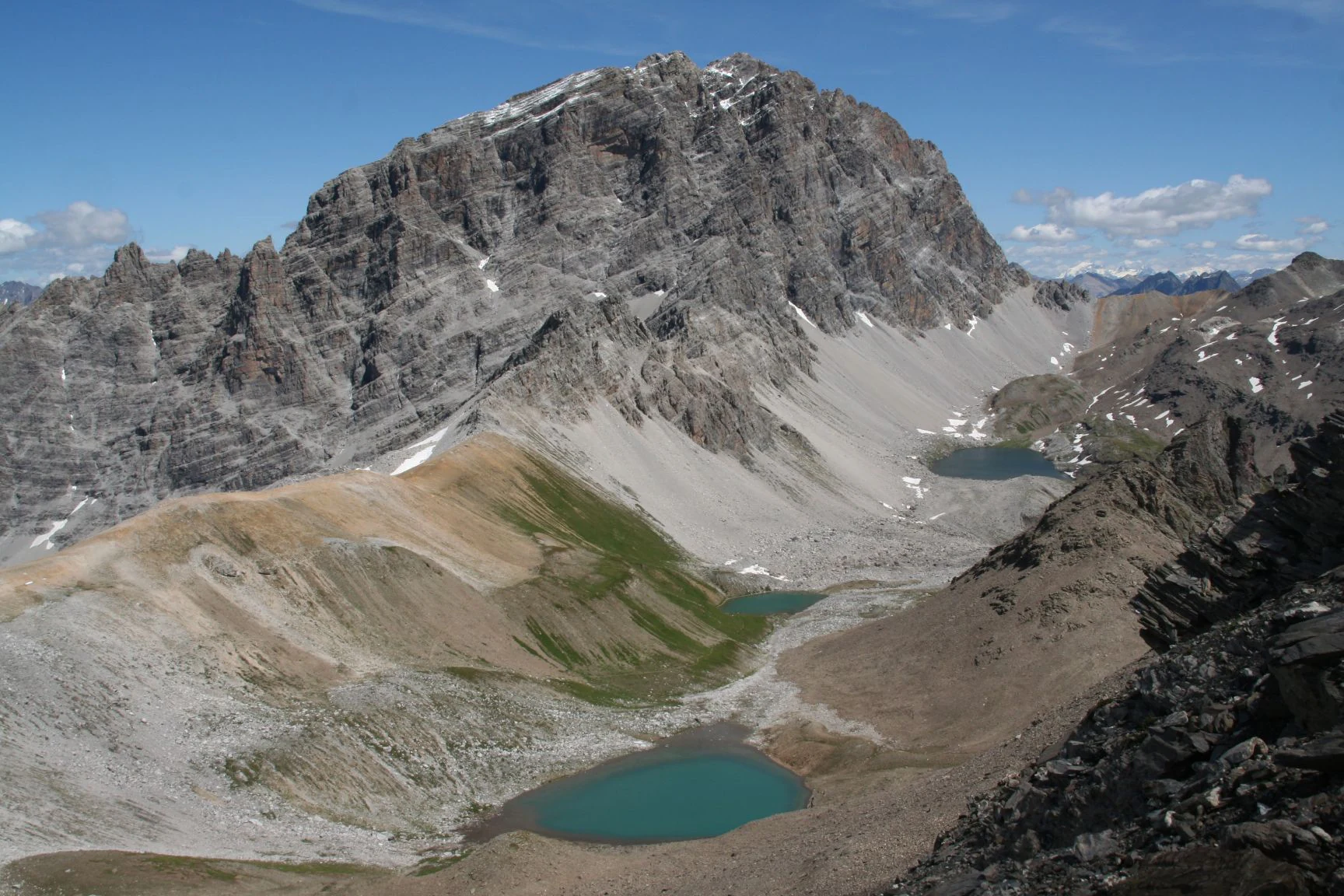 A wide, high-angle mountain view showing the craggy, grey limestone peak of Piz Ela in the center, with small turquoise glacial lakes and scree slopes visible in the foreground and midground.