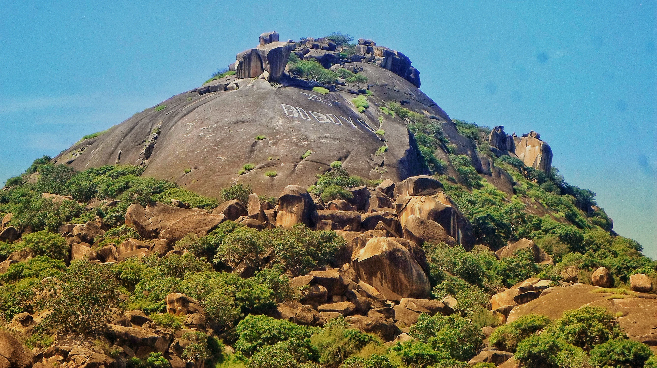 A large, rounded granite dome peak known as Mount Boboyo rising above surrounding dry shrubland and scattered green trees.