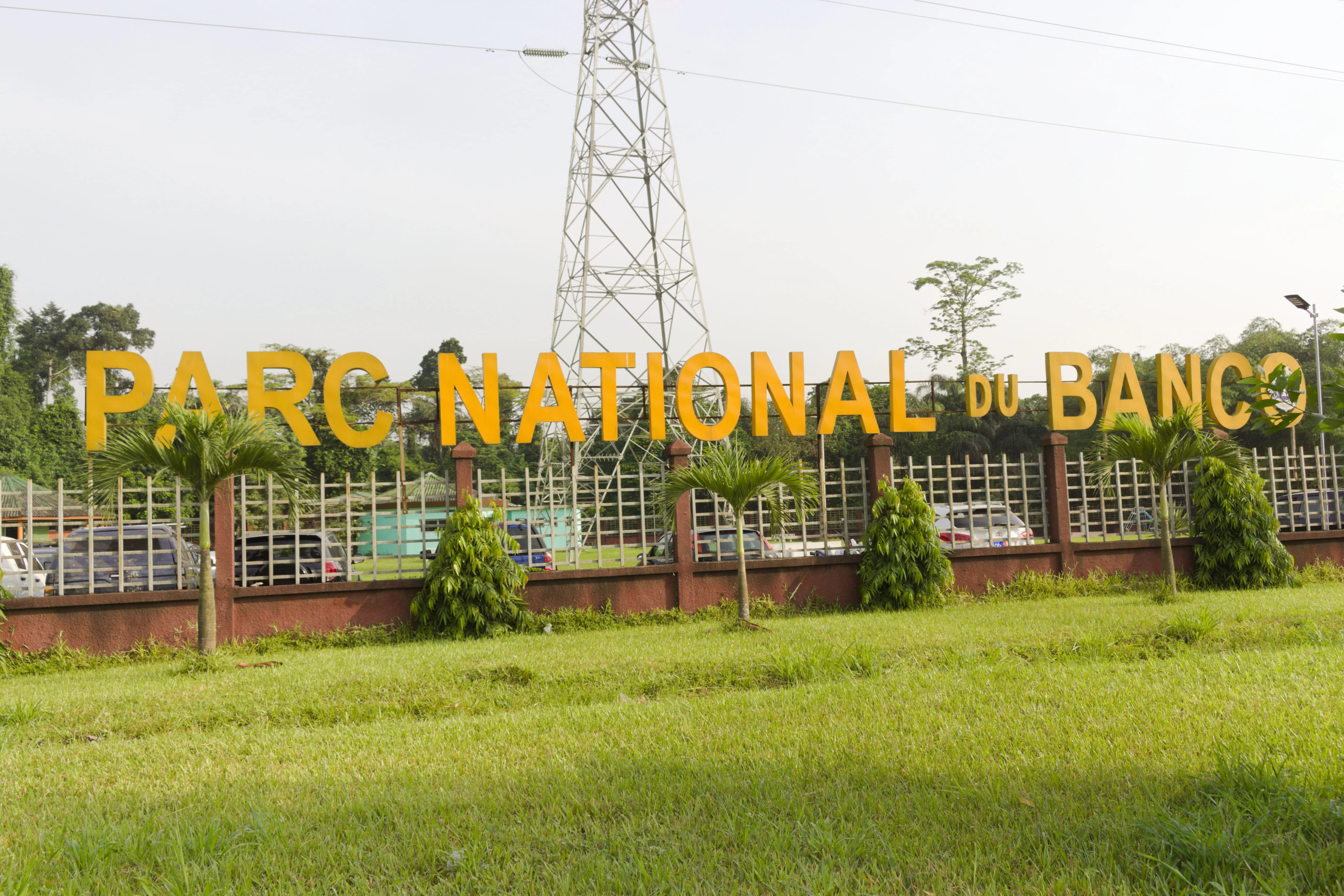 A large yellow sign reading PARC NATIONAL DU BANCO mounted on a gated metal fence, with green trees and a tall electrical transmission tower in the background.