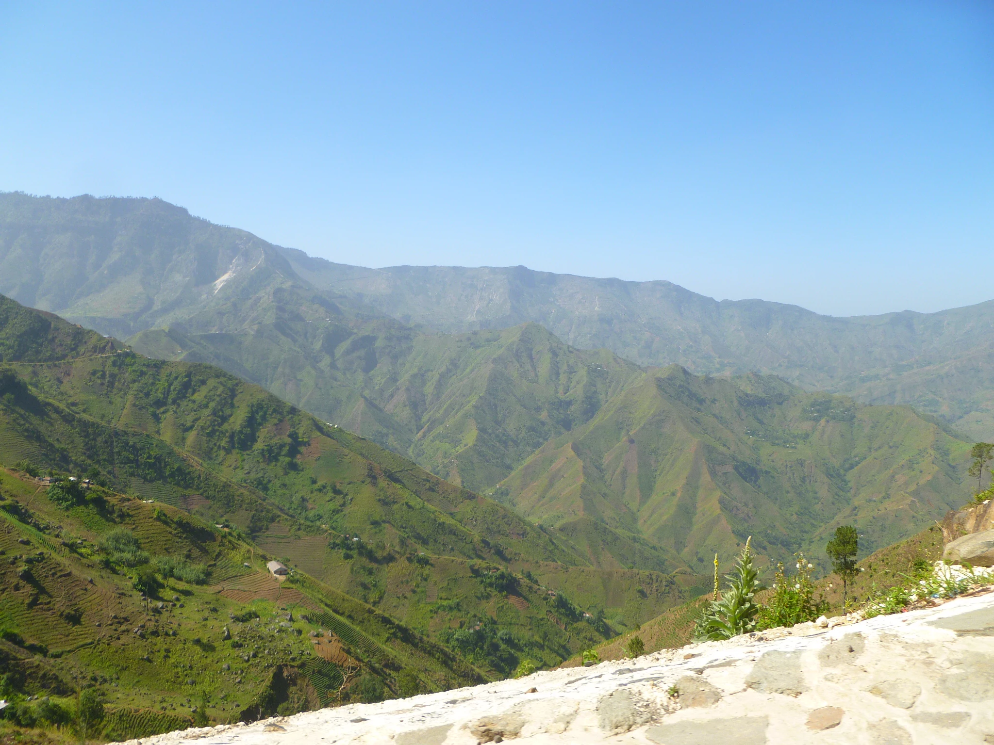 A wide, scenic view looking across a series of deep, green, rugged mountain ridges under a clear blue sky, with a stone-paved edge visible in the immediate foreground.