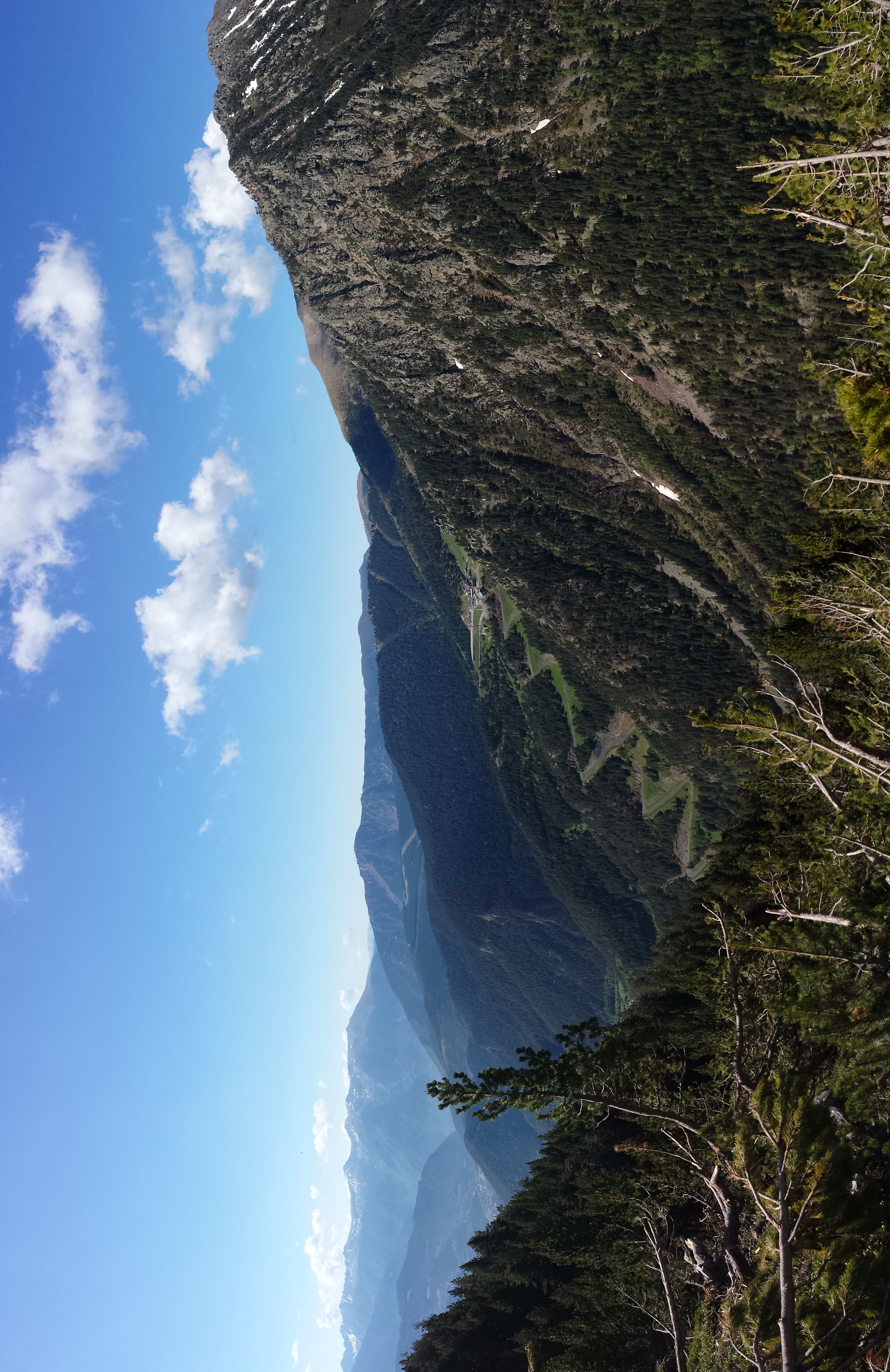 Wide view of mountain valleys with forested slopes under a clear blue sky, featuring distant peaks and scattered clouds